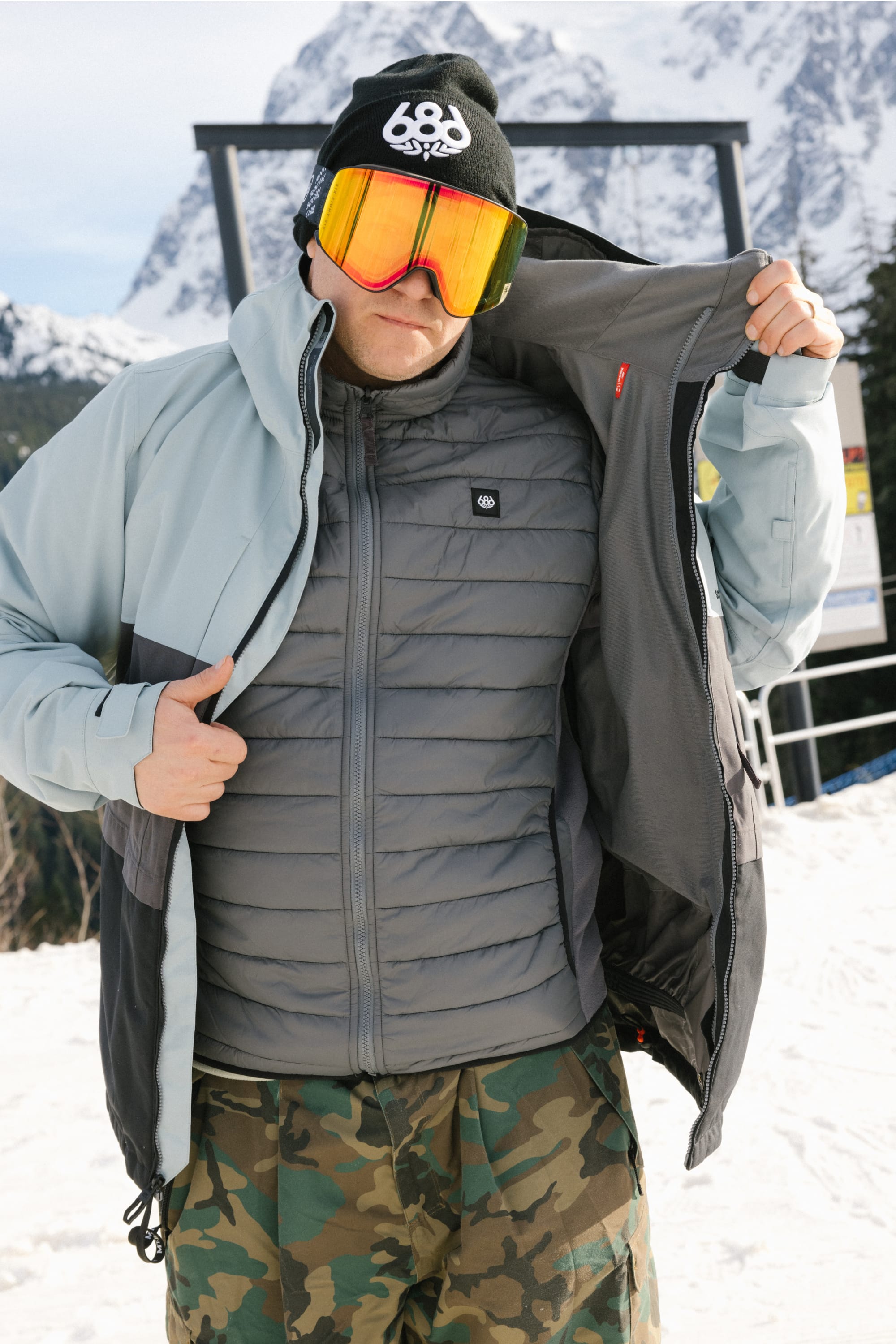 A man wearing a black beanie, orange ski goggles, camouflage pants, and the 686 Men's SMARTY® 3-In-1 Form Jacket by 686 stands on snow with mountains behind him, unzipping his jacket.