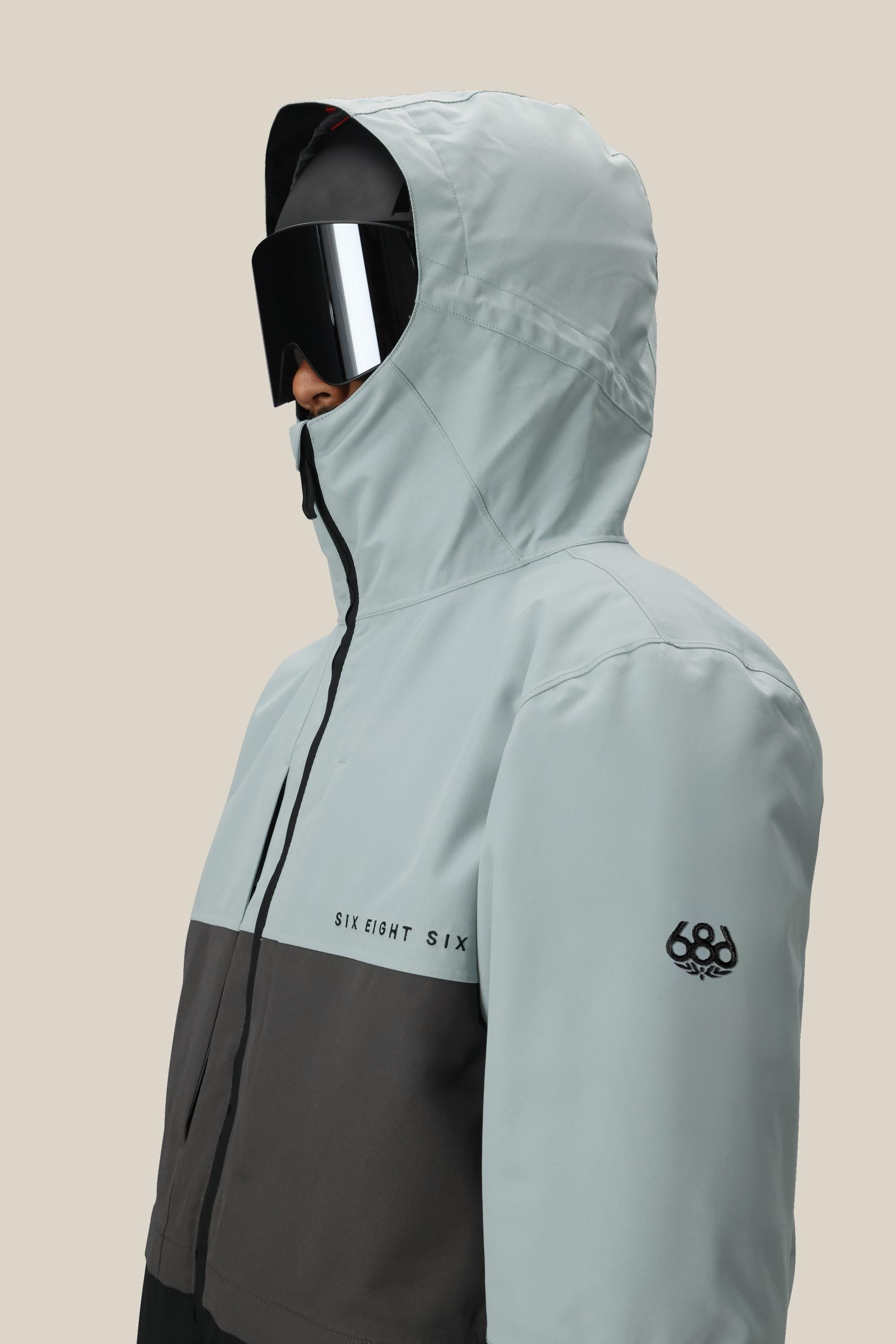 A man models the 686 Men's SMARTY® 3-In-1 Form Jacket in light gray and black with SIX EIGHT SIX lettering and sleeve logo, paired with large black ski goggles, against a beige background. This waterproof polyester jacket is both stylish and functional.