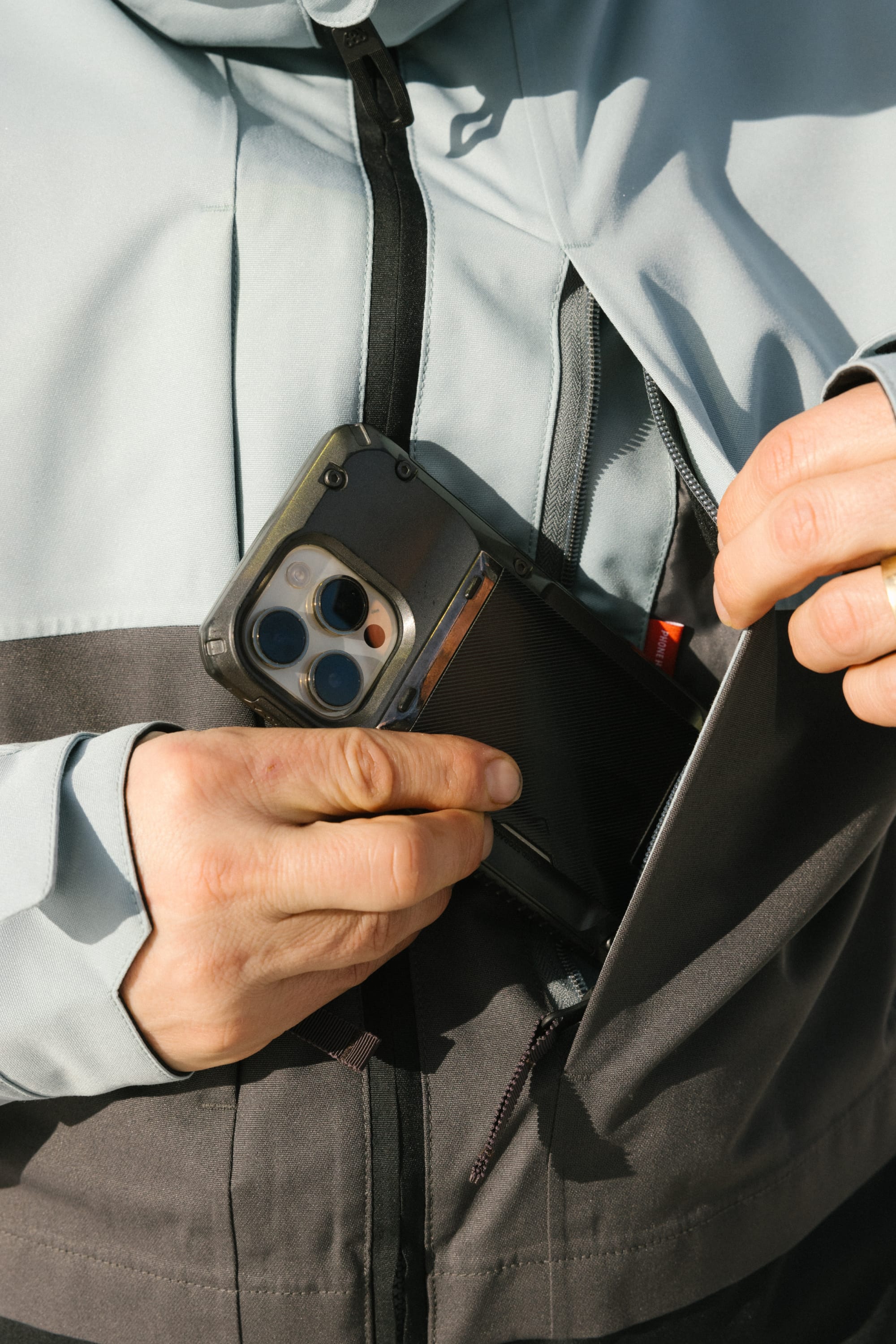 A person wearing the 686 Men's SMARTY® 3-In-1 Form Jacket in light gray and black is placing a smartphone into the outer pocket, with a close-up focus on their hands and phone.