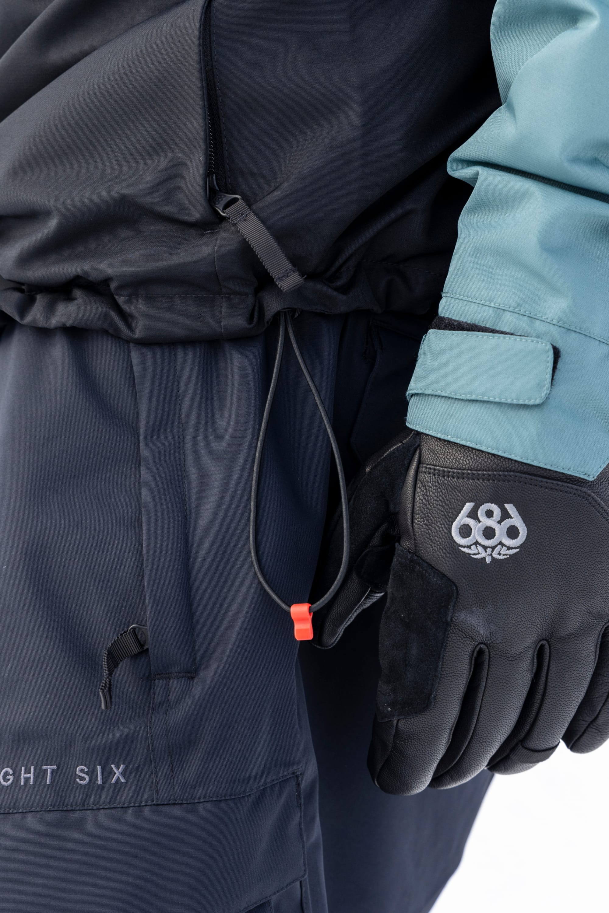 A close-up of a person wearing dark snow pants, black gloves with the 686 logo, and the 686 Men’s SMARTY® 3-In-1 Form Jacket in blue and black, pulling on an elastic cord with a red toggle.