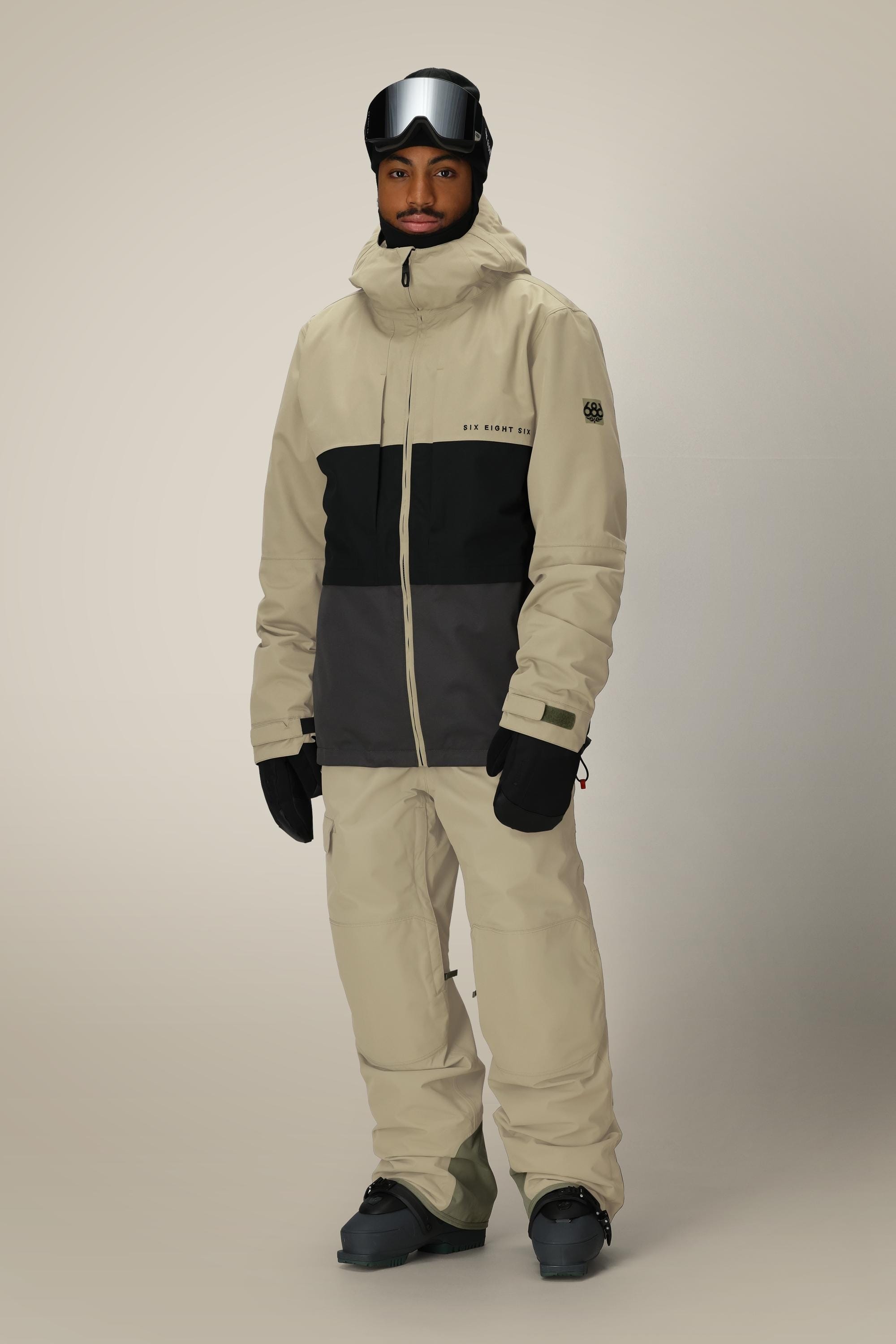 A person wearing the 686 Men's SMARTY® 3-In-1 Form Jacket in beige and black, along with ski gear such as snow pants, gloves, boots, and ski goggles, stands against a plain light background.