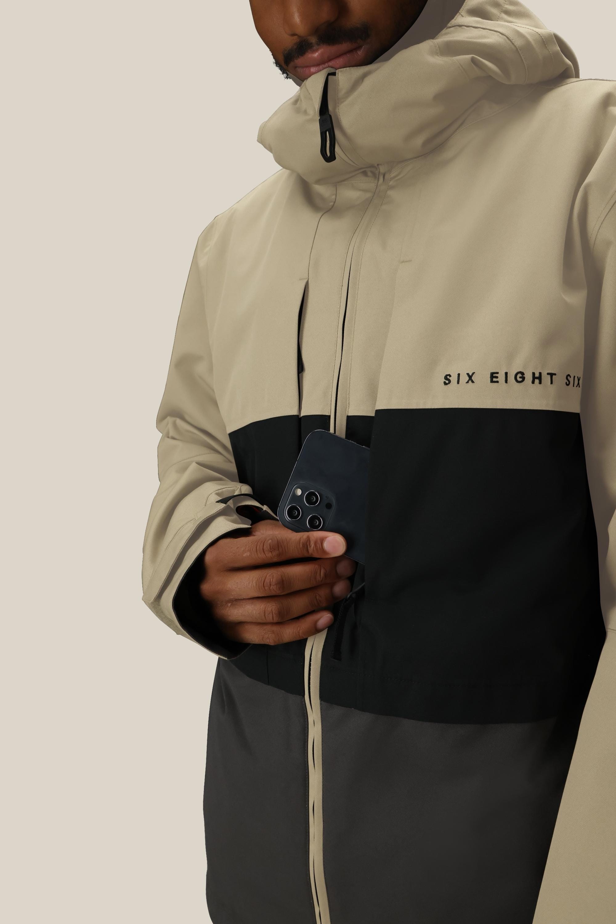 A person wearing the 686 Men's SMARTY® 3-In-1 Form Jacket in beige and black places a black smartphone into their chest pocket, with only the lower half of their face and upper torso visible.