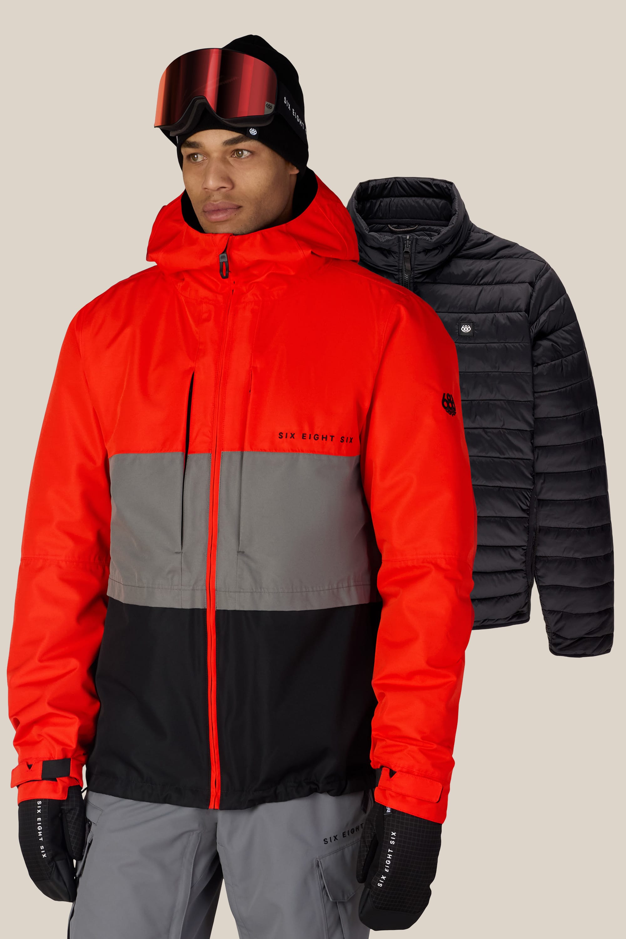 A man is wearing the 686 Men’s SMARTY® 3-In-1 Form Jacket by 686, paired with gray pants, black gloves, a black beanie, and ski goggles. Behind him, a black puffer jacket is displayed on a plain background.