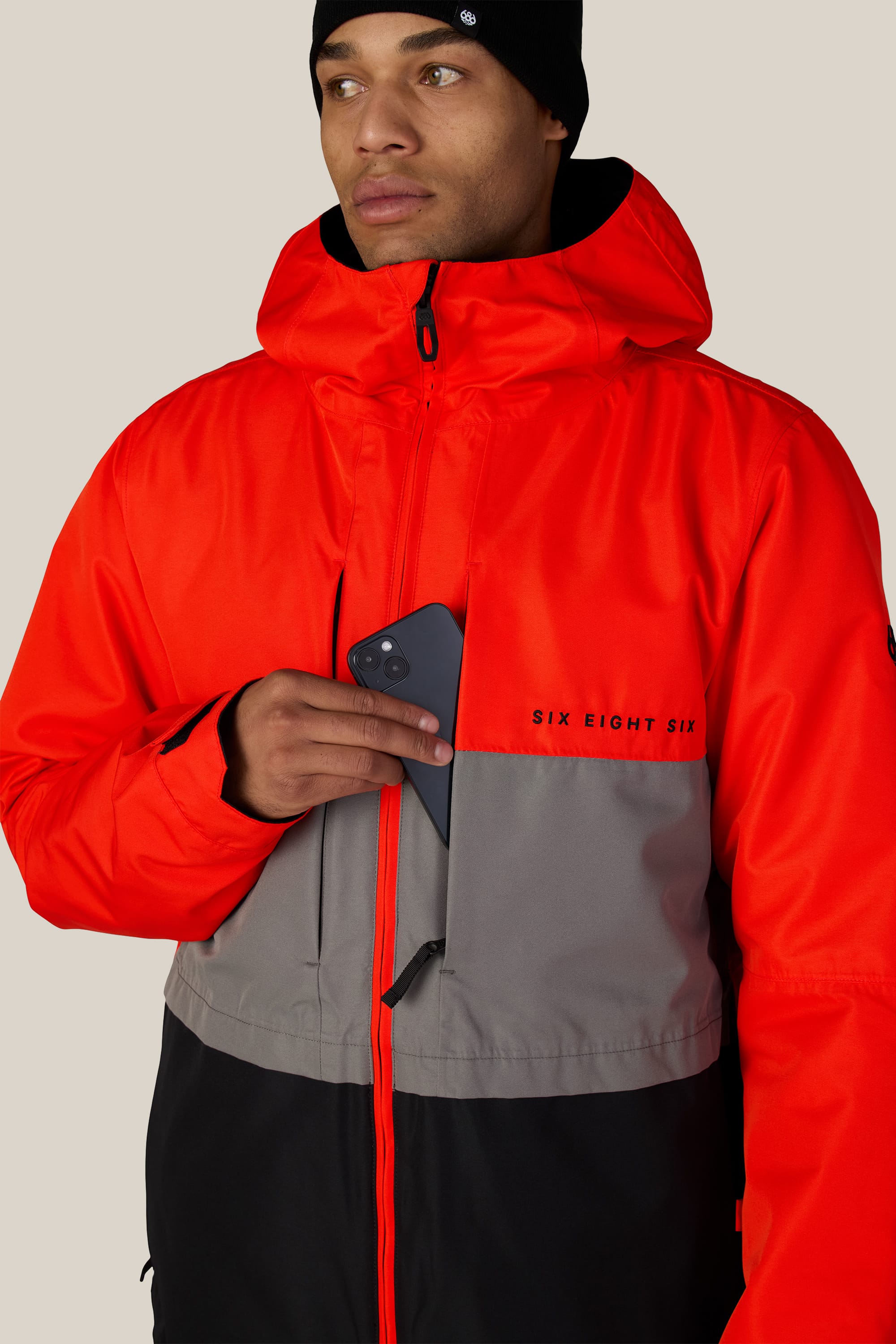 A person wearing the 686 Men’s SMARTY® 3-In-1 Form Jacket in red, gray, and black, featuring the 686 brand name, holds a black smartphone and wears a black beanie while looking to the side against a neutral background.