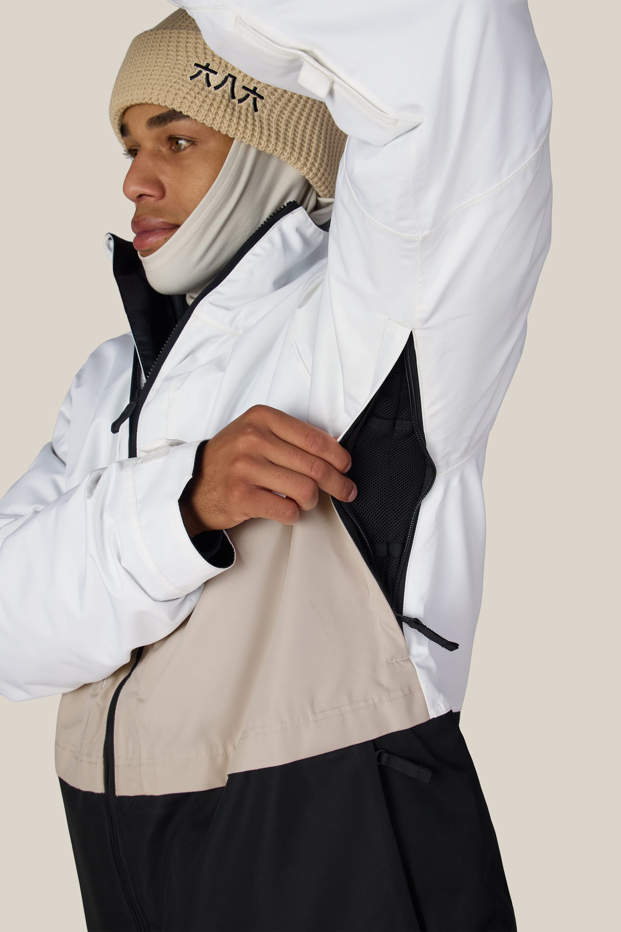 Wearing the 686 Men’s SMARTY® 3-In-1 Form Jacket in white and beige along with a tan beanie, a person opens the underarm zipper to showcase the jacket’s ventilation feature. The background is plain and light-colored.
