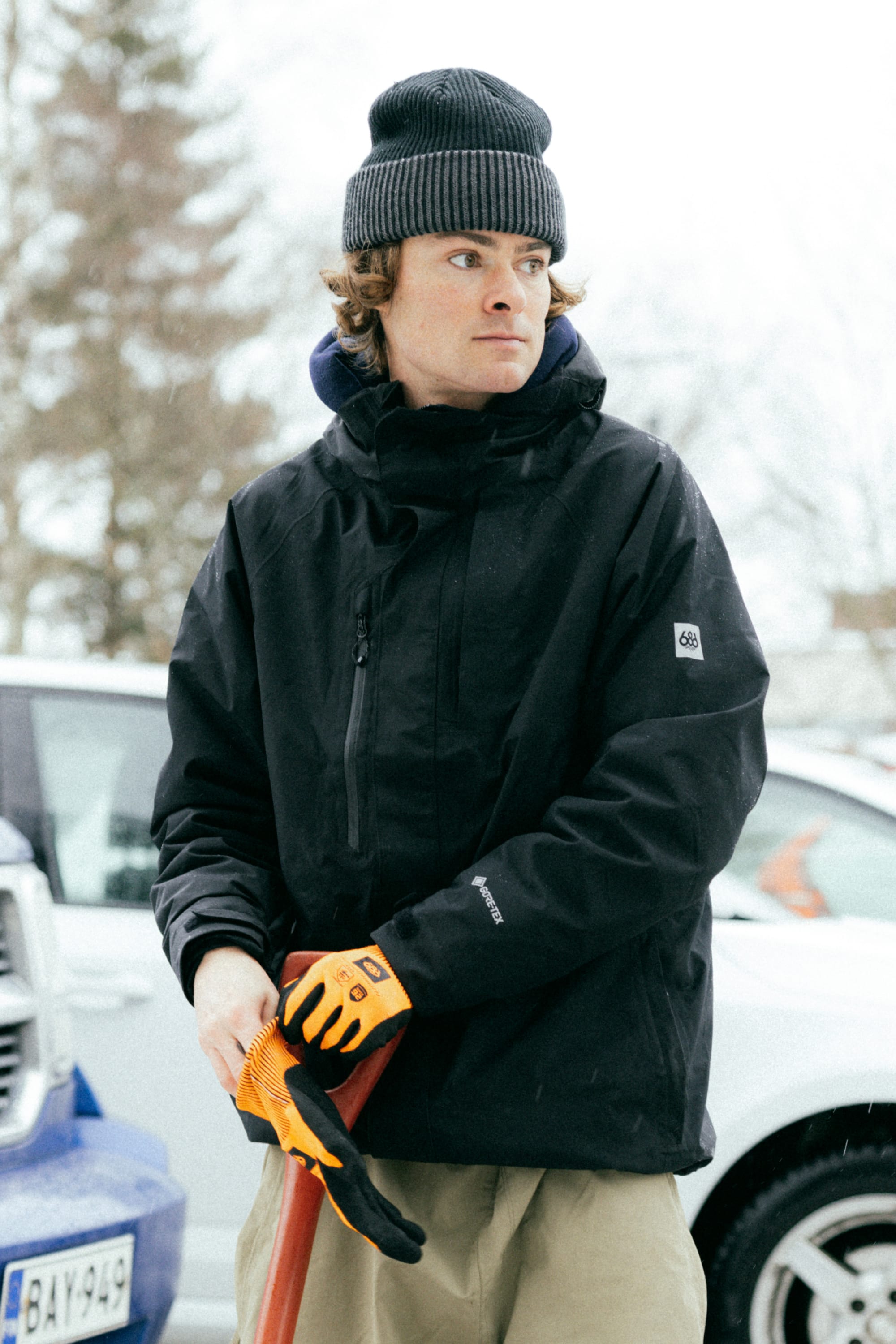 A person wearing the 686 Men’s GORE-TEX® Core Shell Jacket by 686, along with a gray beanie and orange gloves, stands outdoors in winter holding a shovel, with cars and snowy trees in the background.