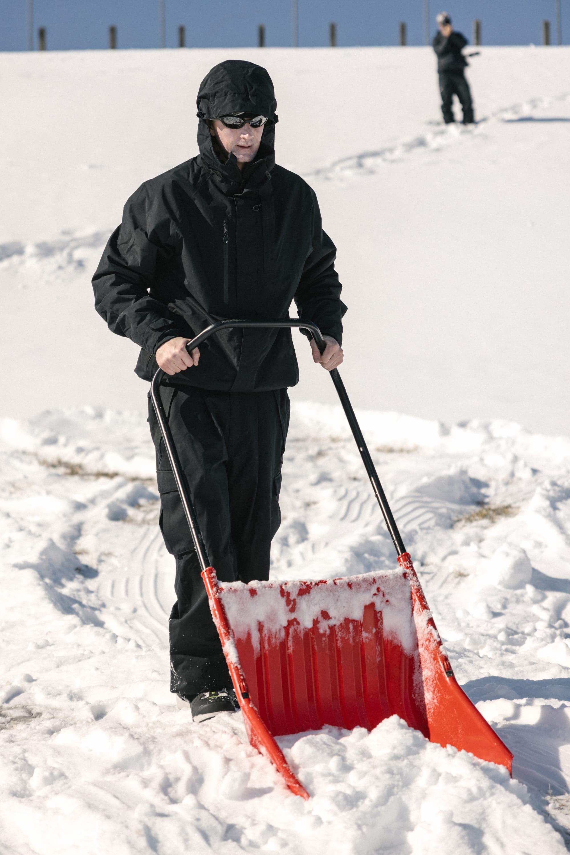 Wearing the 686 Men’s GORE-TEX® Core Shell Jacket, a person uses a red snow pusher to clear snow while another stands in the snowy background.
