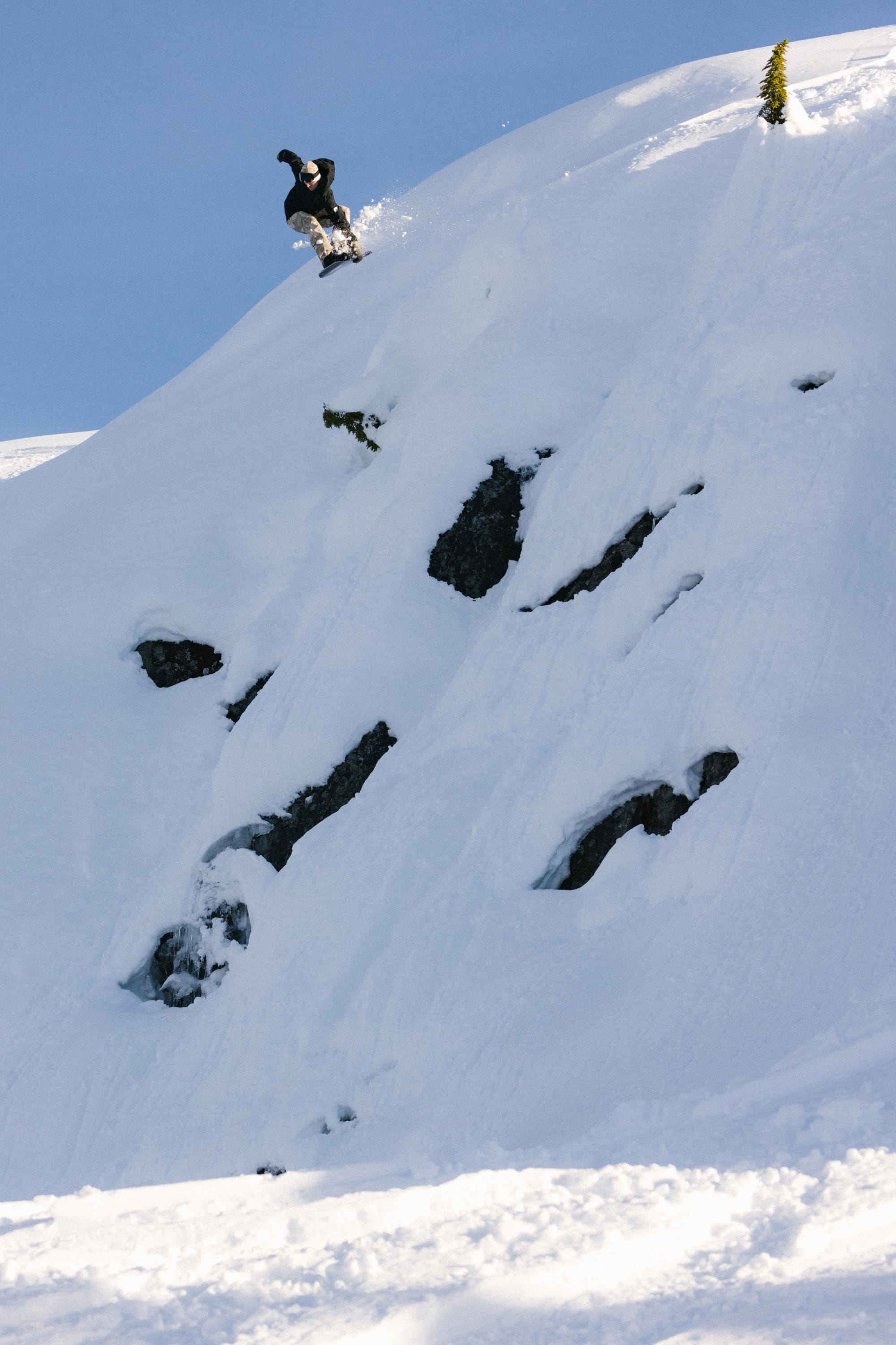 A snowboarder wearing the 686 Mens GORE-TEX Core Shell Jacket rides down a steep, snow-covered mountain slope with exposed rocks under a clear blue sky.