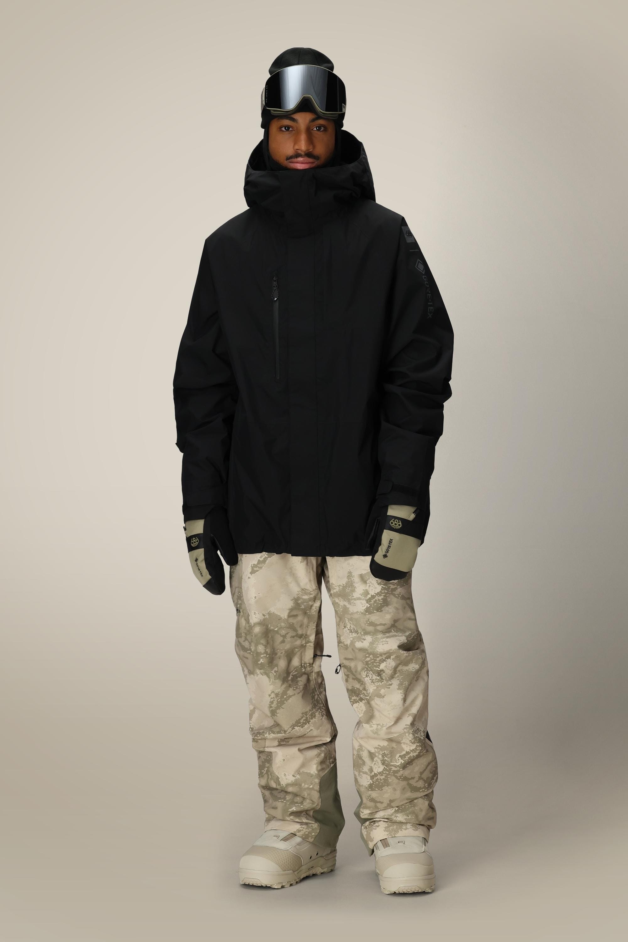 A person stands facing forward against a plain light background, wearing the 686 Men’s GORE-TEX® Core Shell Jacket in black, paired with beige camouflage snow pants, gloves, white boots, and ski goggles.