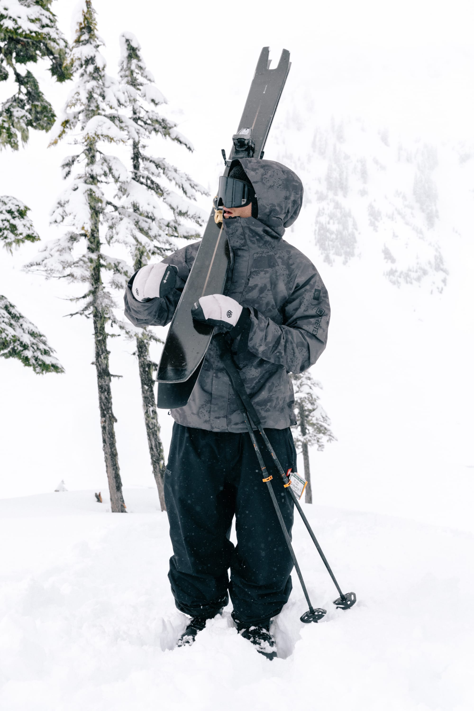 Wearing the 686 Men’s GORE-TEX® Core Shell Jacket, a person stands in the snow holding skis and ski poles, surrounded by snow-covered trees and a wintry landscape.