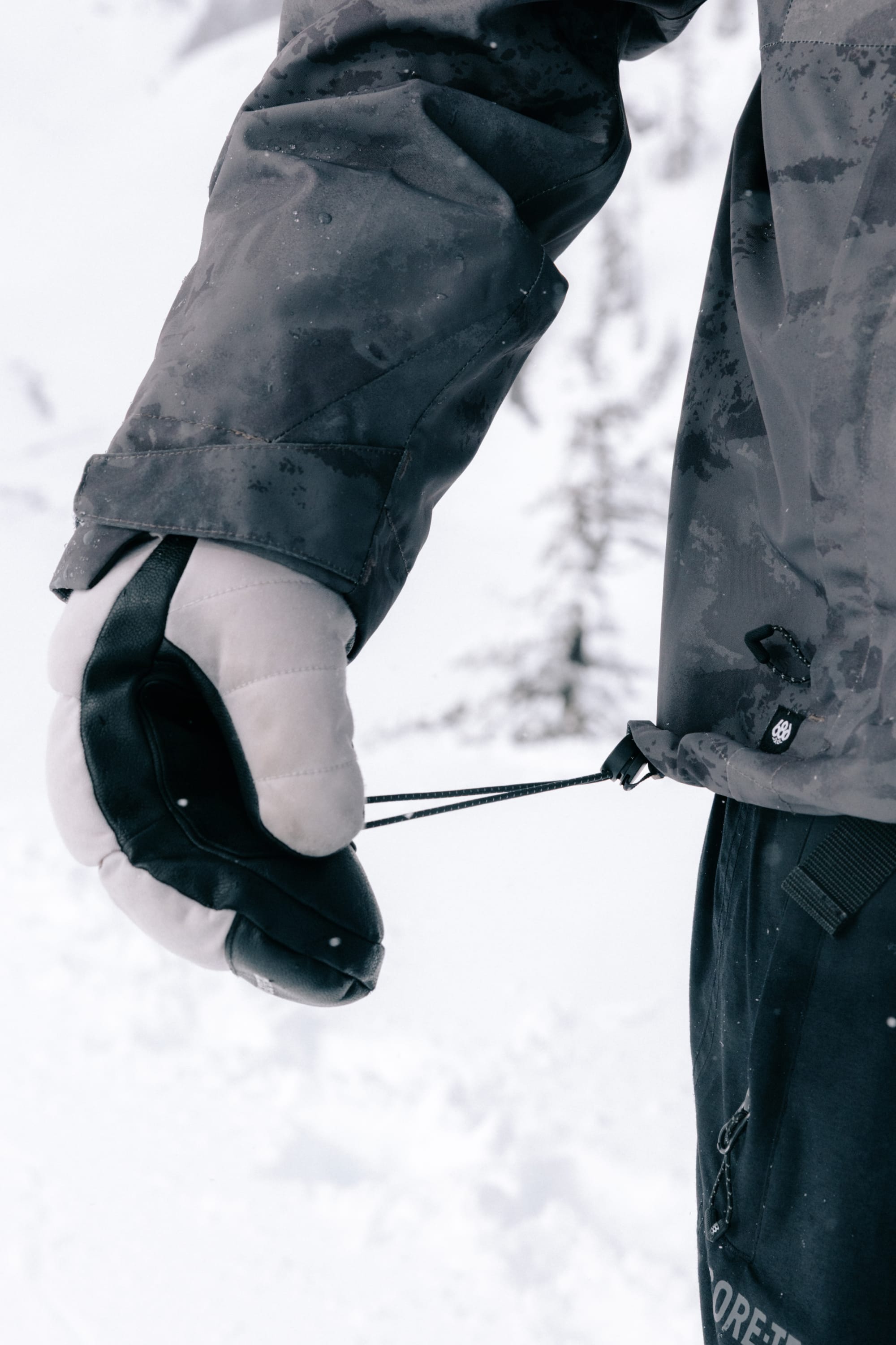 A person wearing the 686 Men’s GORE-TEX® Core Shell Jacket and a white glove holds a black object attached to a drawstring in a snowy outdoor setting.