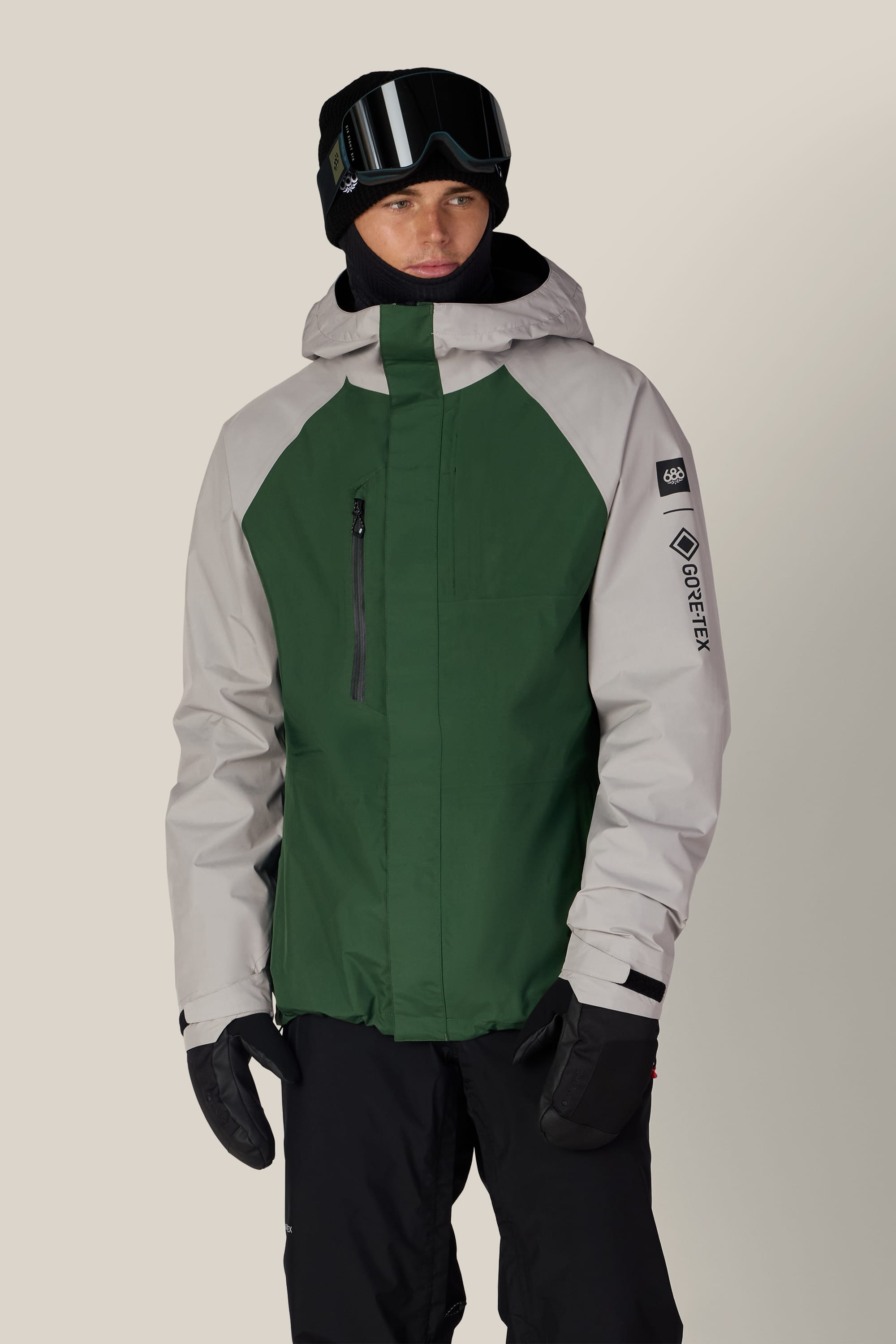 Wearing the 686 Men’s GORE-TEX® Core Shell Jacket by 686, paired with black snow pants, mittens, and ski goggles, a person poses against a plain background.