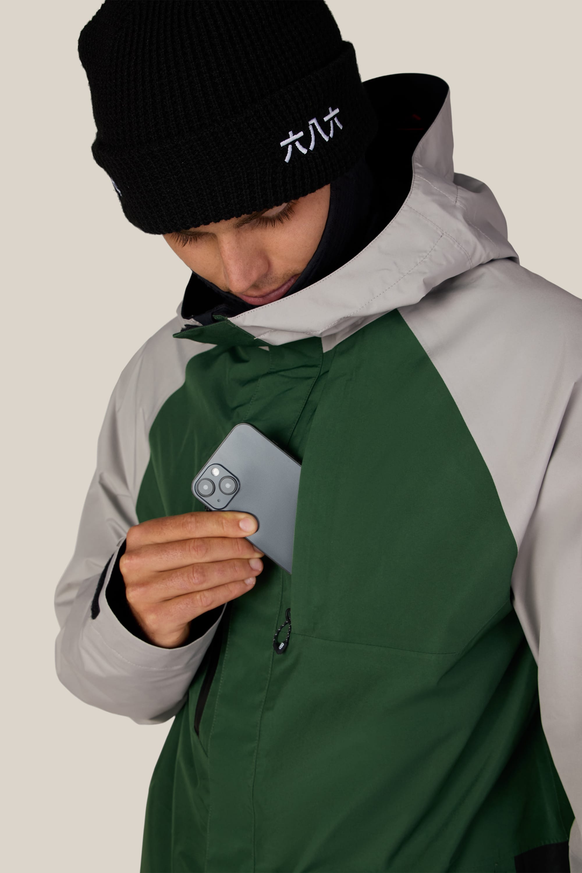 A person wearing the 686 Men’s GORE-TEX® Core Shell Jacket in green and gray places a smartphone into its zippered chest pocket. They are also wearing a black beanie with white embroidered text.