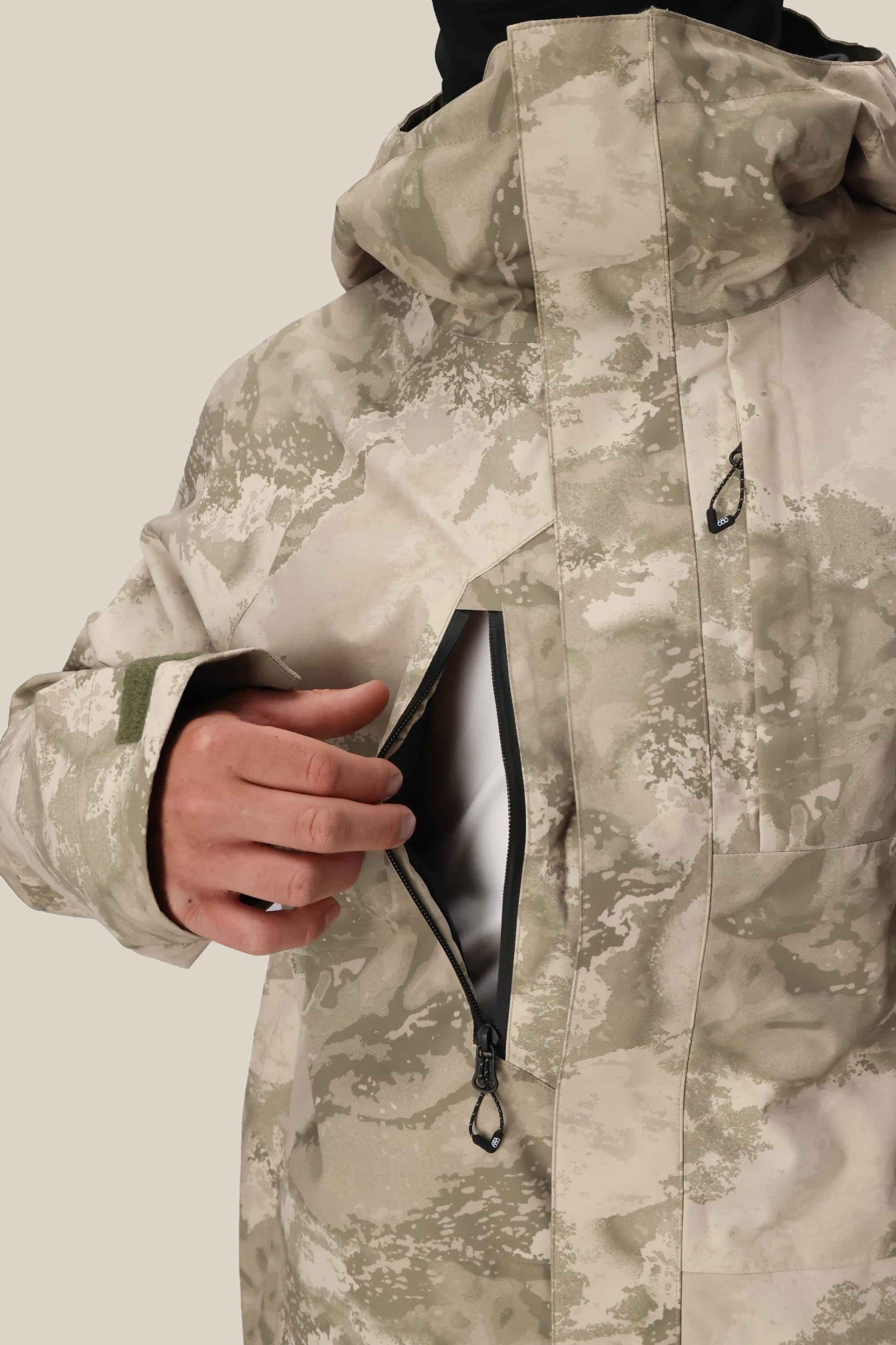 A person, partially visible, unzips the chest pocket of a 686 Mens GORE-TEX Core Shell Jacket in light beige and gray. The jacket features a high collar, hood, Velcro cuffs, and is fully waterproof.