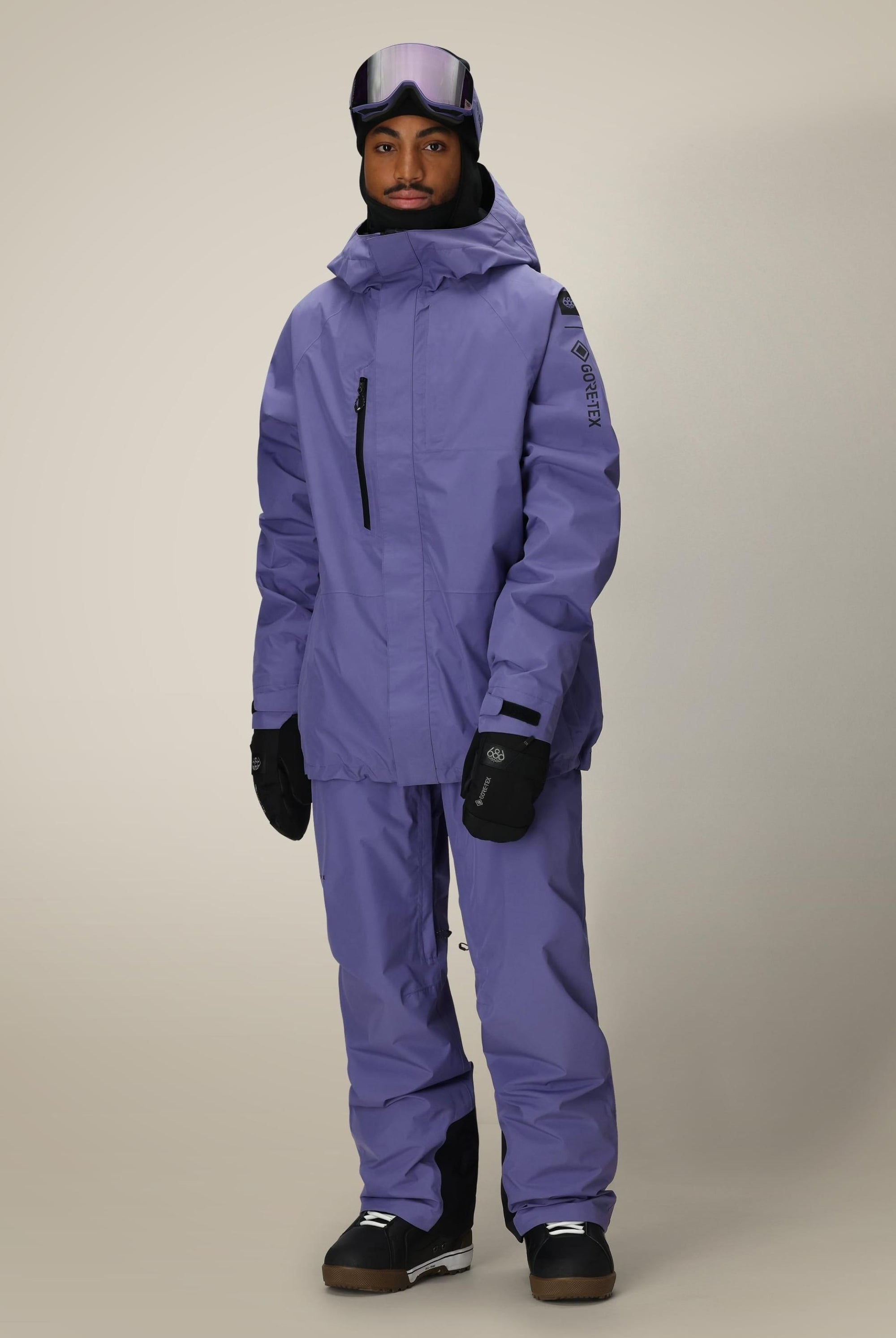 A person stands facing forward against a light background, wearing the 686 Mens GORE-TEX Core Shell Jacket in purple, matching pants, black gloves and boots, with ski goggles resting on their forehead.