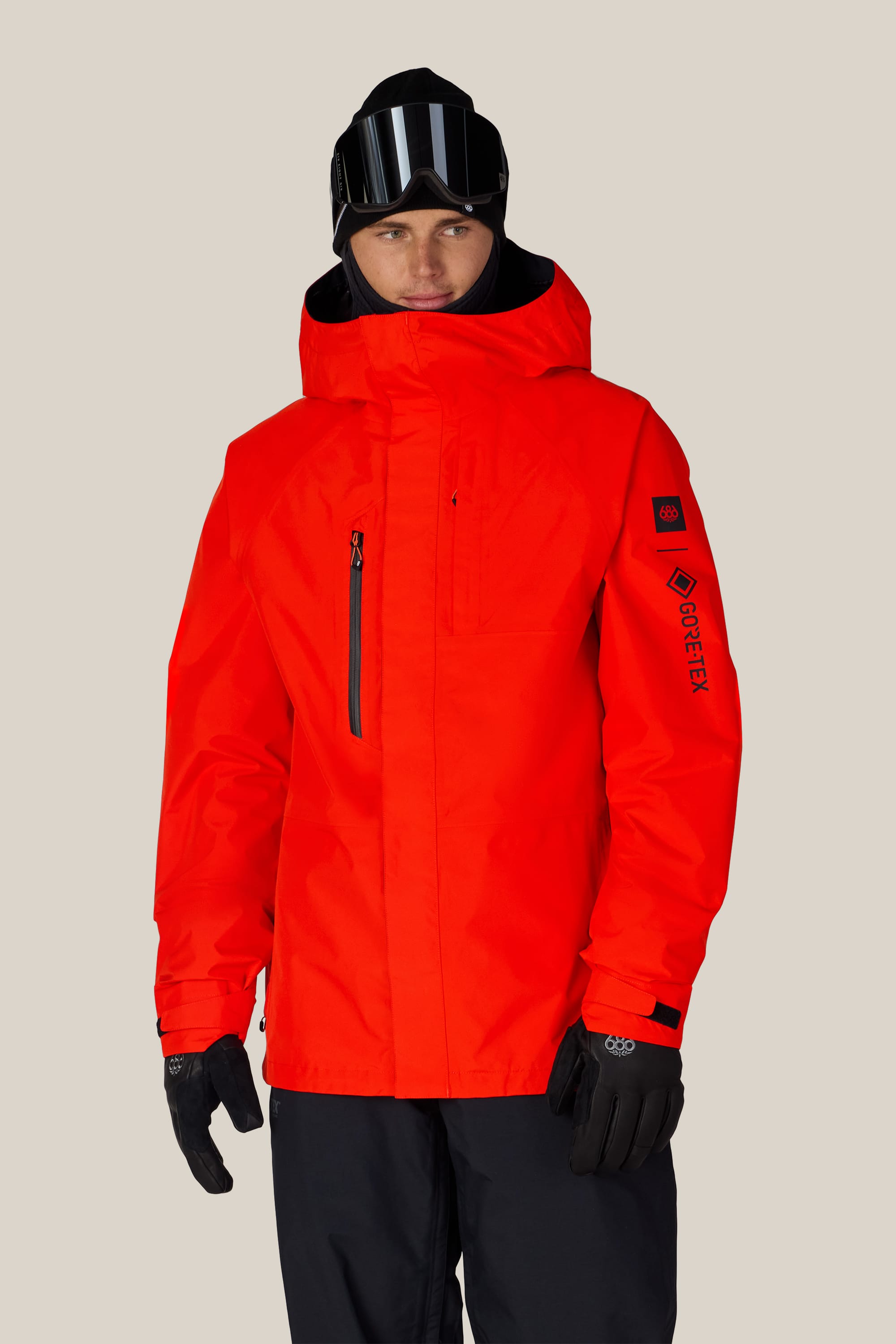 Wearing the 686 Men’s GORE-TEX® Core Shell Jacket in bright red, along with black gloves, black pants, and a black beanie topped with ski goggles, the individual stands against a plain light background.