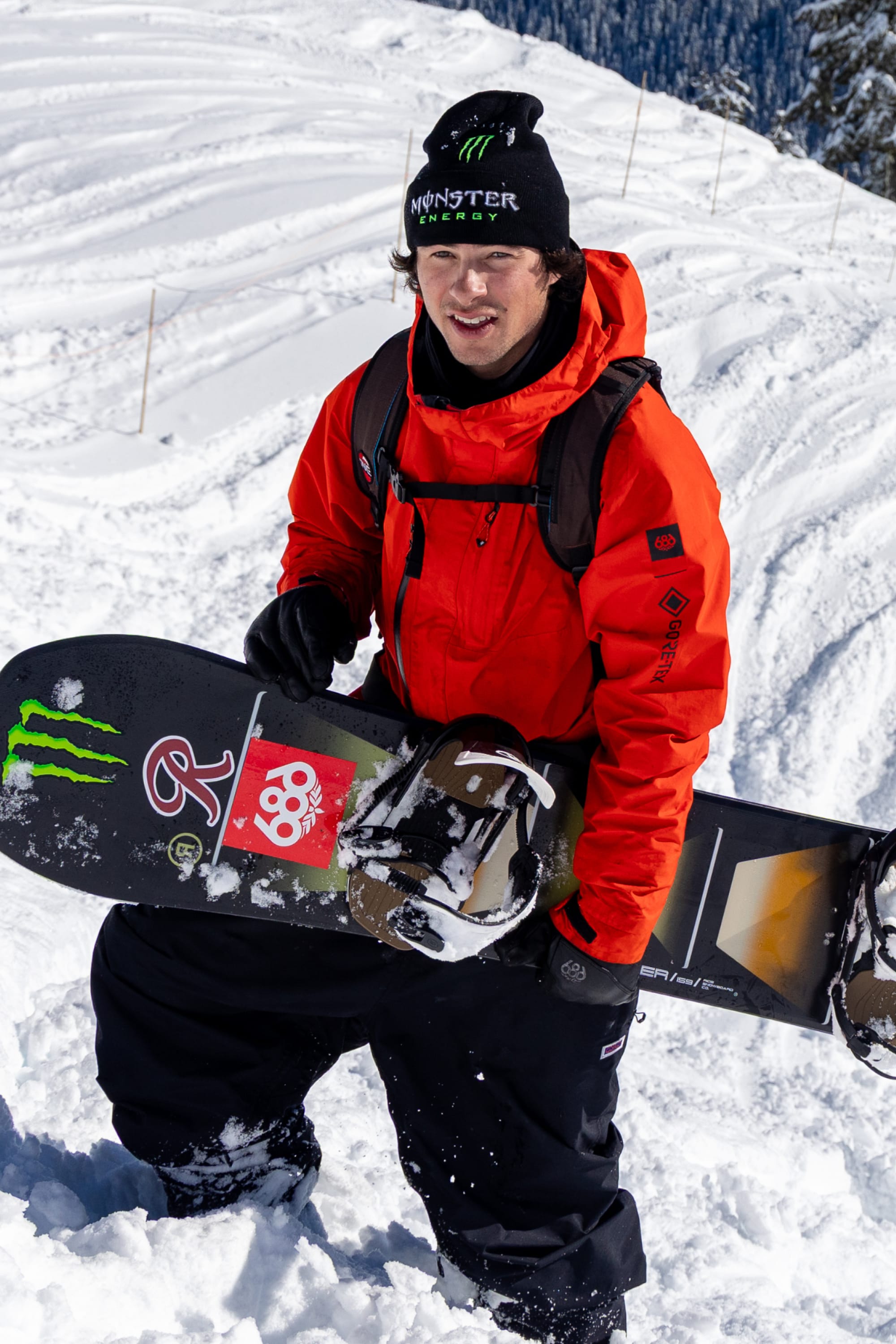 Wearing the 686 Men’s GORE-TEX® Core Shell Jacket in bright red, a snowboarder stands on snowy terrain in black pants and a black hat, holding a snowboard adorned with stickers. Snow-covered mountains and trees provide a scenic backdrop.