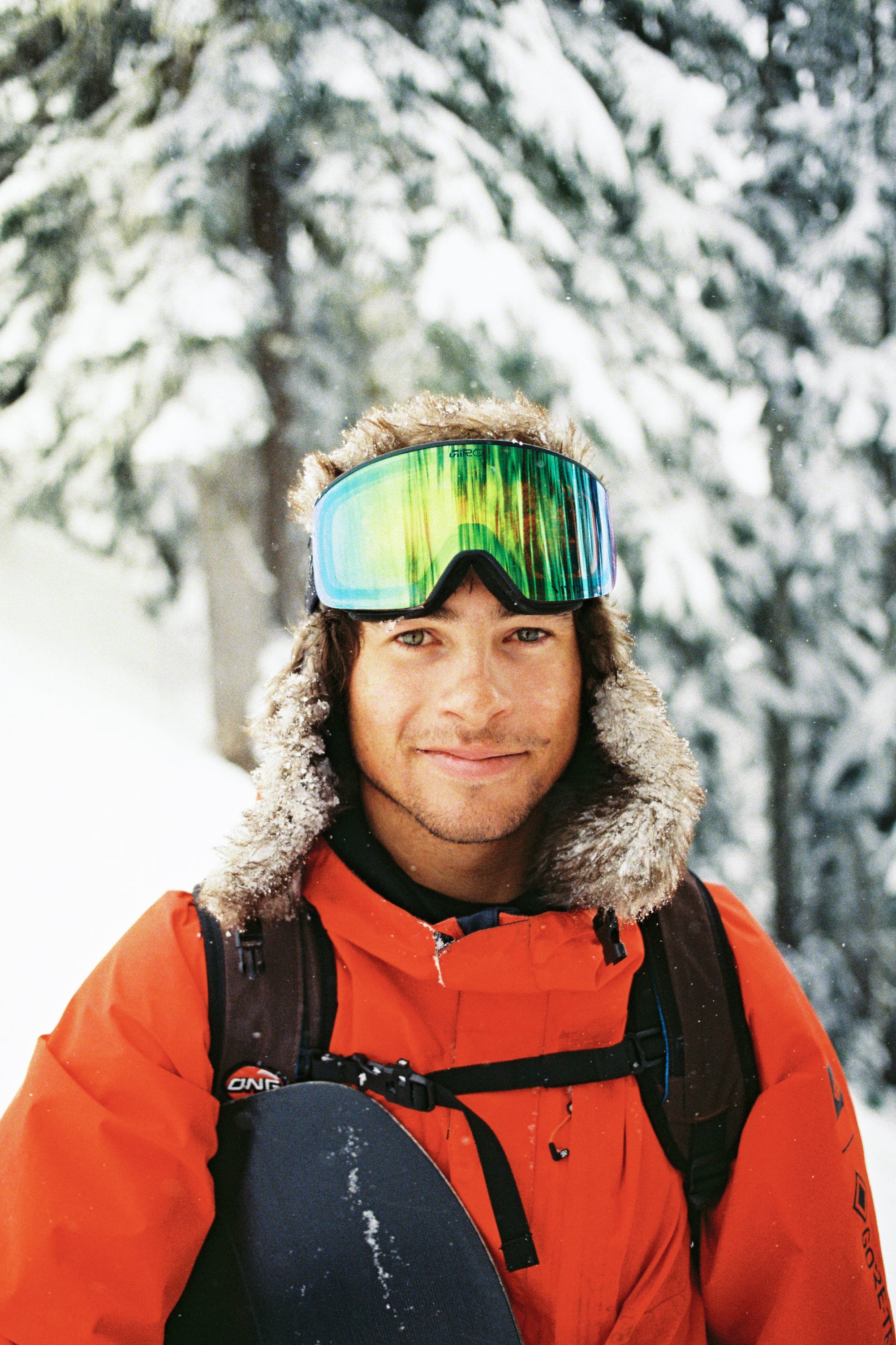 A person in a 686 Men’s GORE-TEX® Core Shell Jacket, fur-lined hat, and reflective ski goggles smiles outdoors in a snowy, forested area.