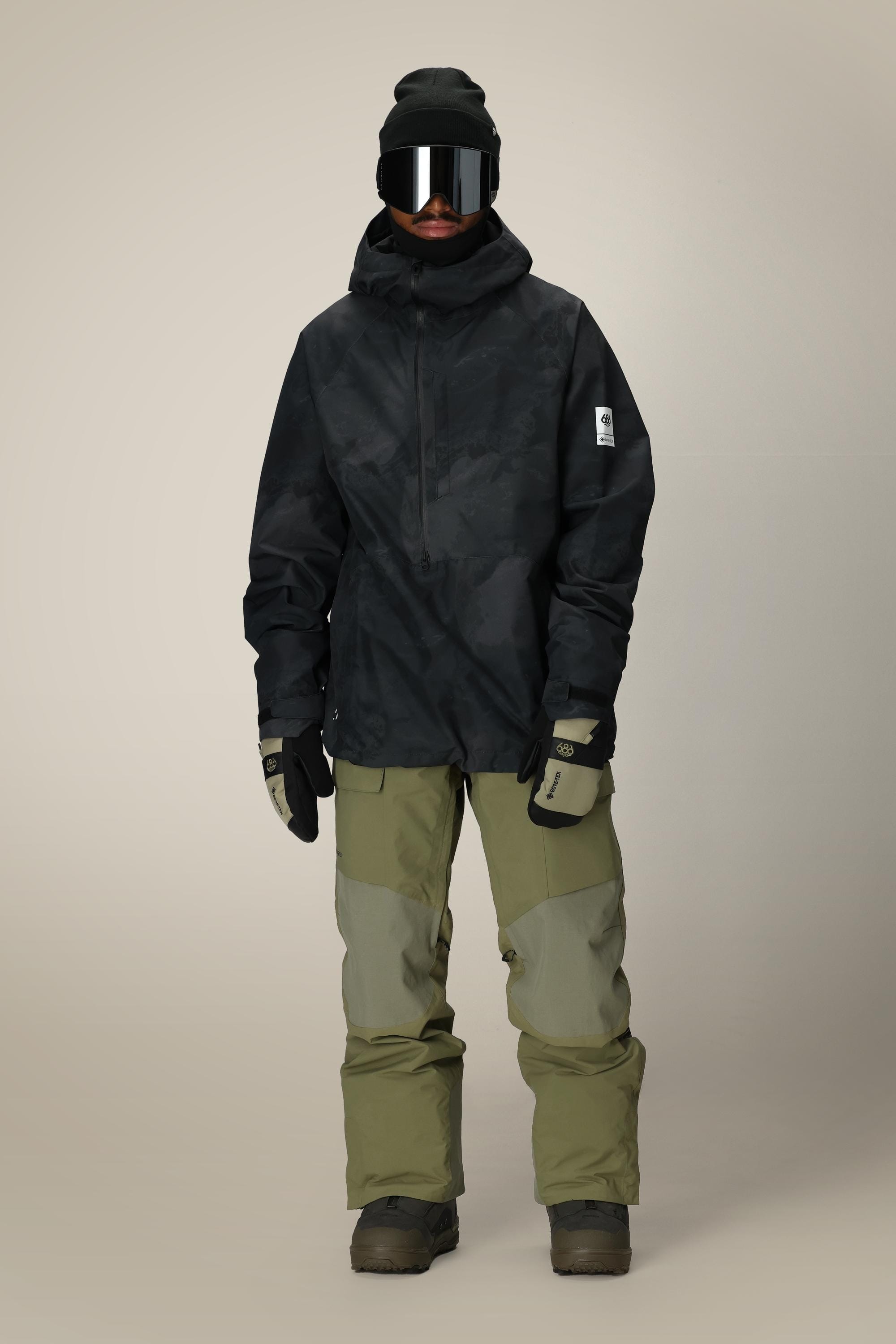 A person wears the 686 Mens GORE-TEX Fragment Shell Anorak, olive green snow pants, gloves, dark boots, a beanie, and ski goggles while standing against a plain light background.