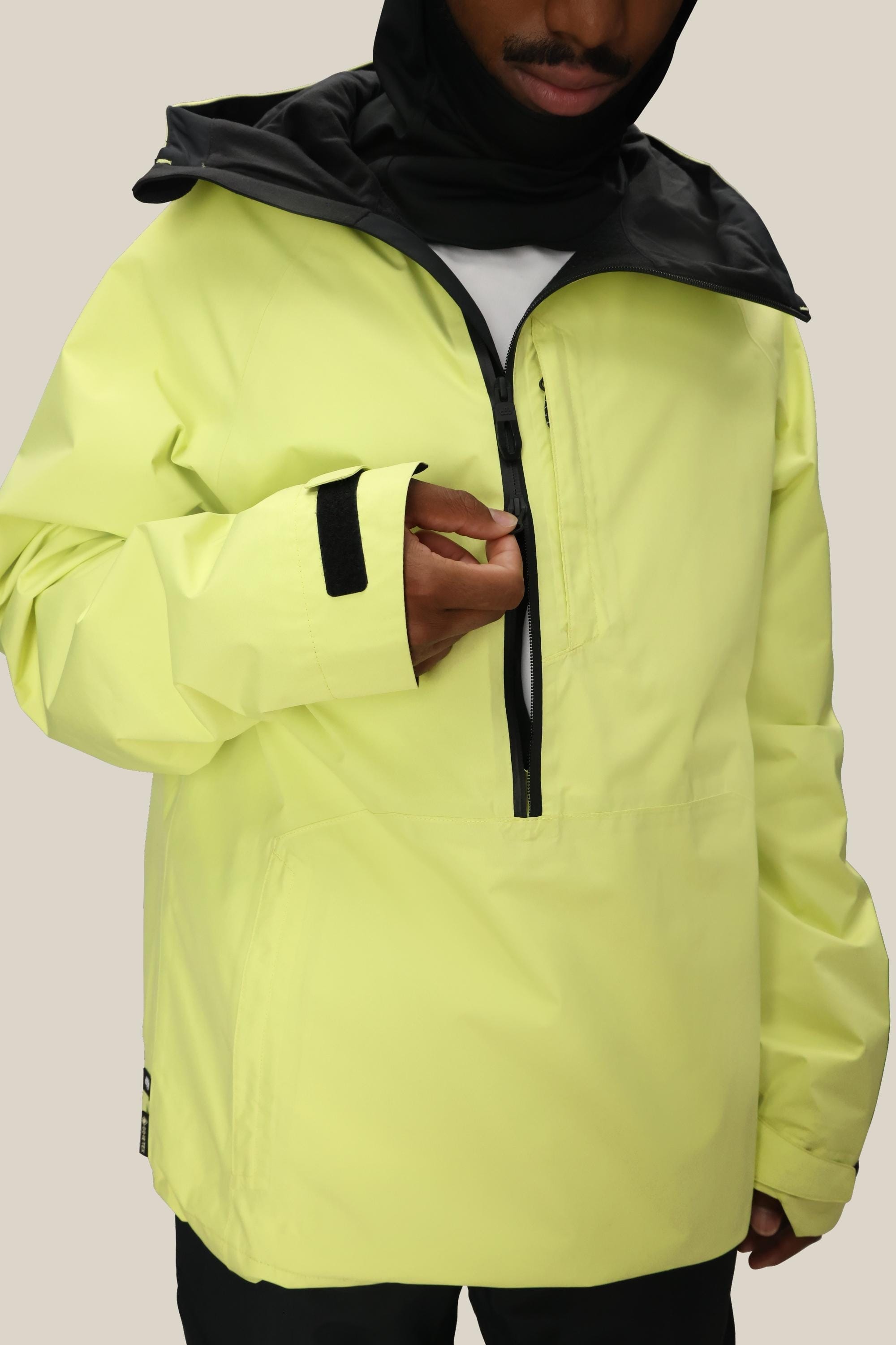 A person wears the 686 Mens GORE-TEX Fragment Shell Anorak in light yellow with black accents, unzipping the chest pocket. The 686 polyester waterproof jacket features a high collar, front zipper, and adjustable cuffs. Face partially visible.