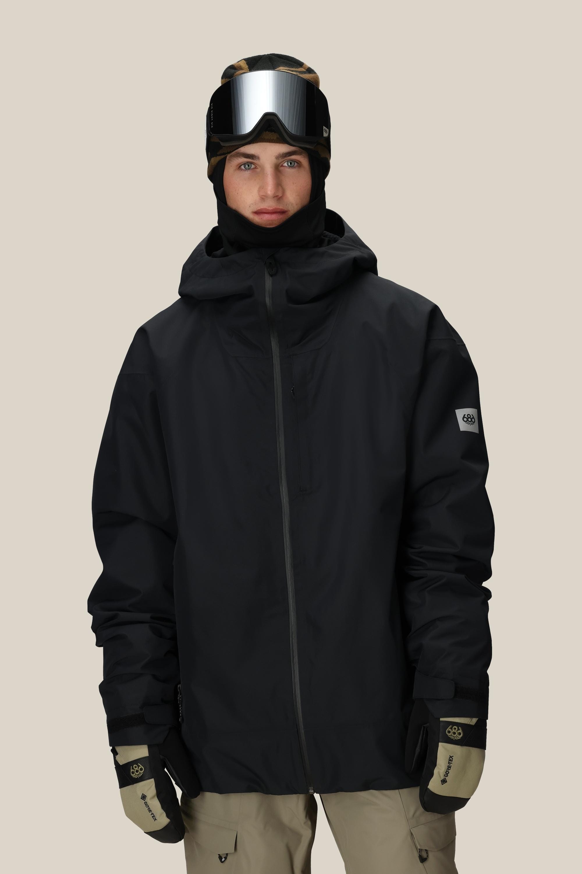 A person stands facing forward against a plain light background wearing the 686 Mens Gateway Shell Jacket with a black hood, beige ski gloves, tan pants, a black beanie, and ski goggles.