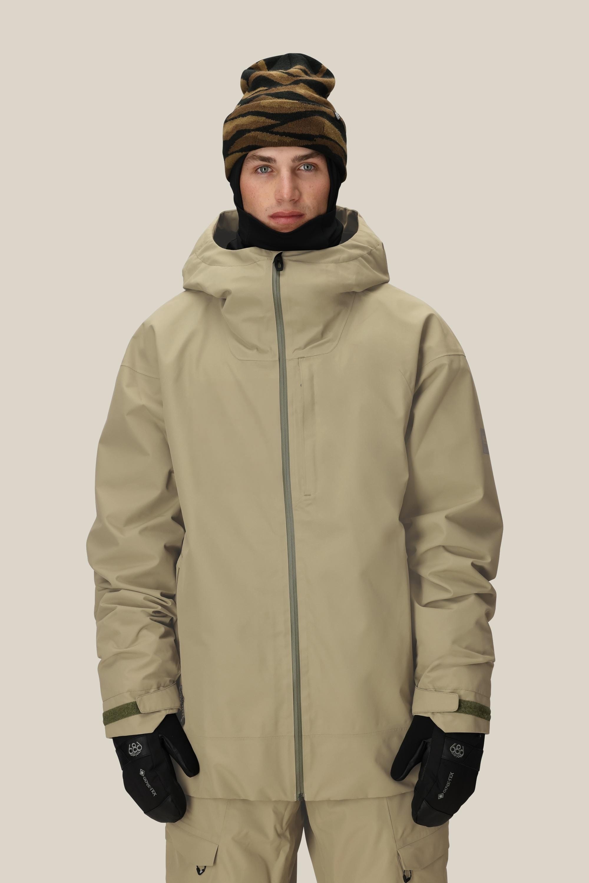 A person wears the 686 Mens Gateway Shell Jacket in tan with matching pants, black gloves, and a black and brown patterned beanie, standing facing forward against a plain light background.