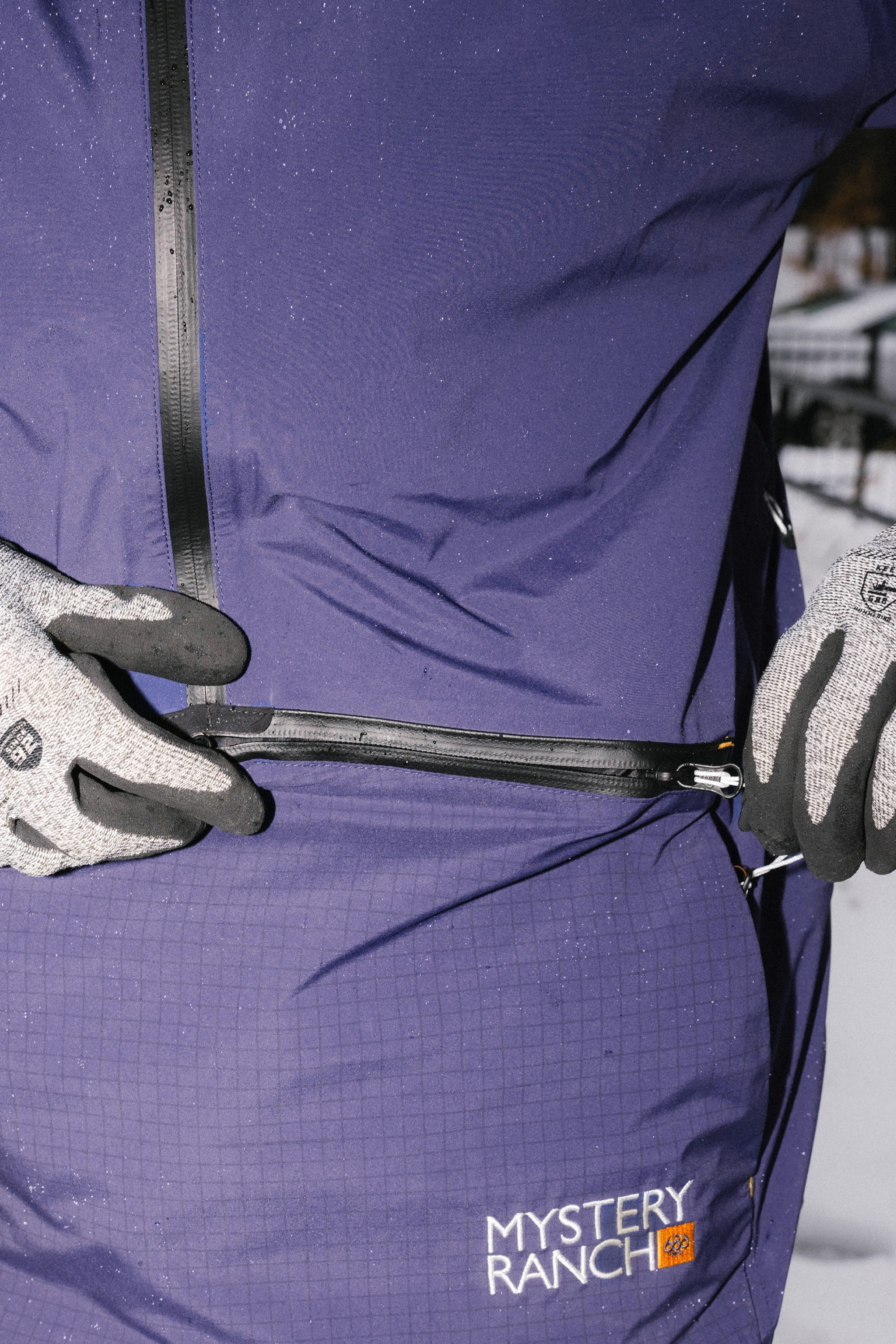 A person wearing gray gloves unzips the waterproof zipper of a purple 686 Men's Mystery Ranch Qi 3L Shell Jacket with logo, outdoors in the snow.