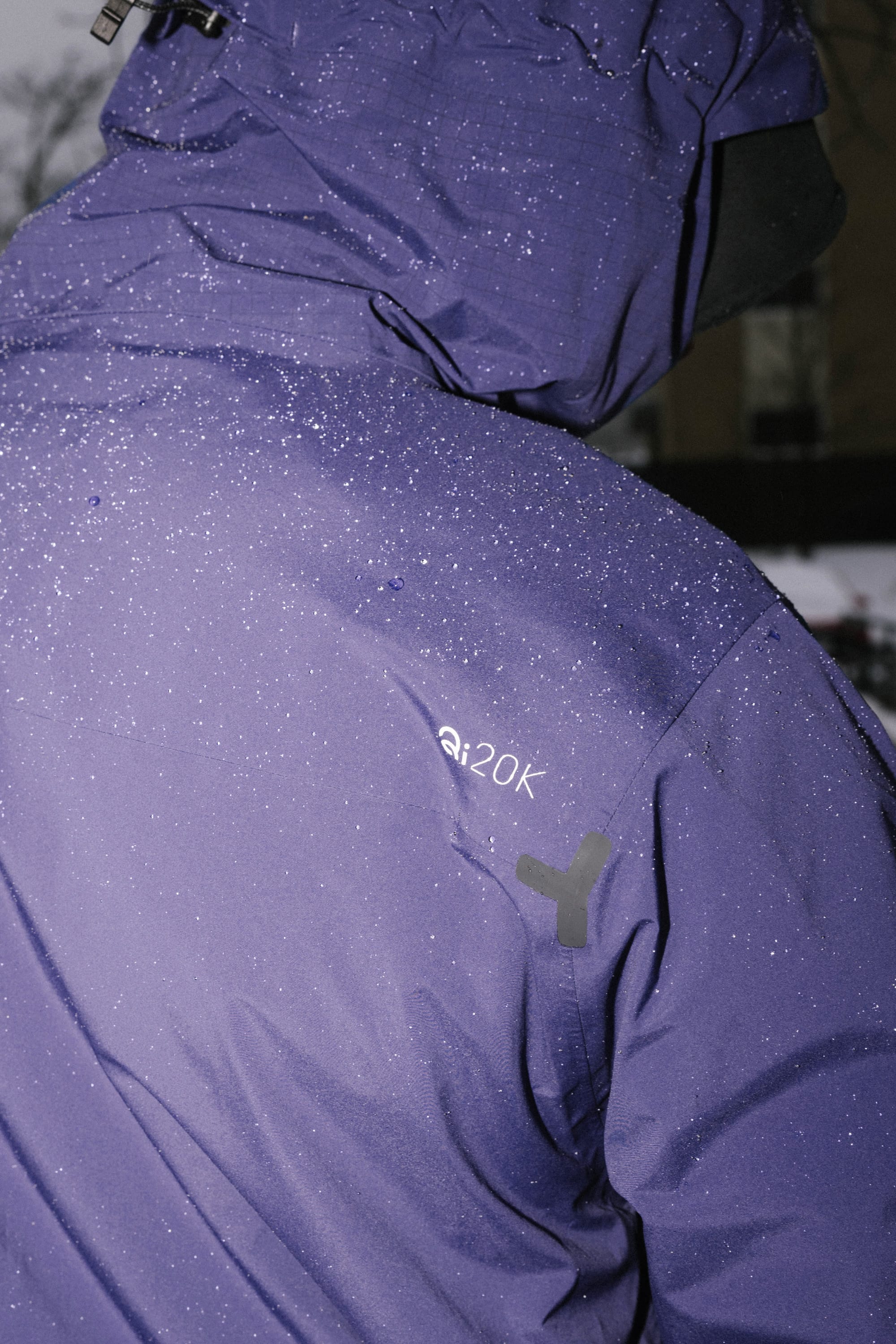 A person wears the 686 Men's Mystery Ranch Qi 3L Shell Jacket, a purple woven polyester waterproof jacket with water droplets on the surface. The hood is up, a "20k" label indicates high resistance, and the blurred background adds emphasis.