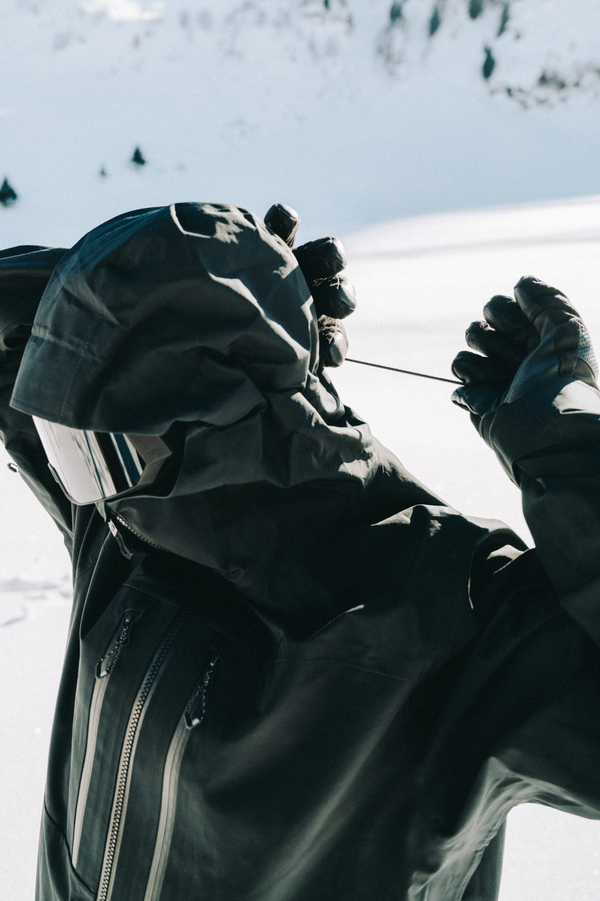 A man in a 686 Men's GORE-TEX 3L ATV Thermagraph® Jacket adjusts the hood drawstring while wearing gloves and ski goggles, standing outdoors in the snow.