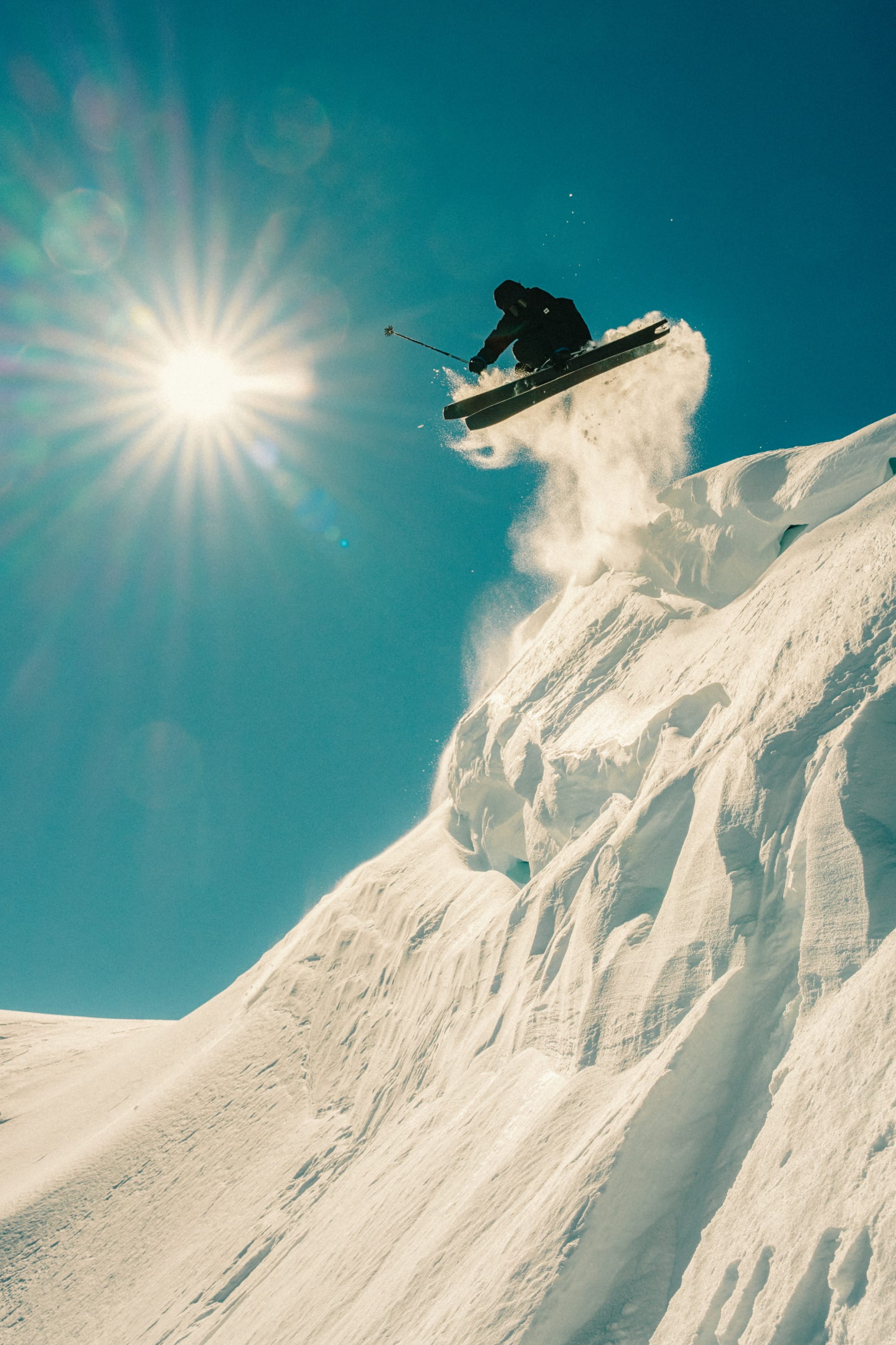 A skier wearing the 686 Men's GORE-TEX 3L ATV Thermagraph® Jacket soars off a snowy cliff under a clear blue sky, sunlight in the background and powdery snow trailing behind.