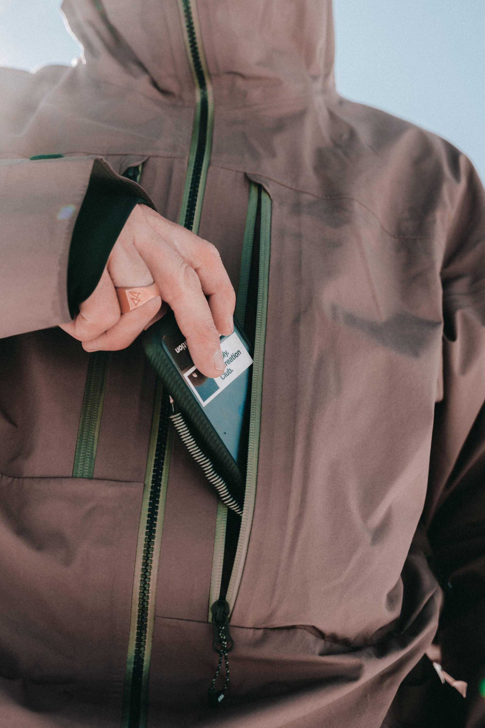 A person in a 686 Men’s GORE-TEX 3L ATV Thermagraph® Jacket slips a small black-and-white device into its zippered chest pocket, with the focus on their hand and the pocket.