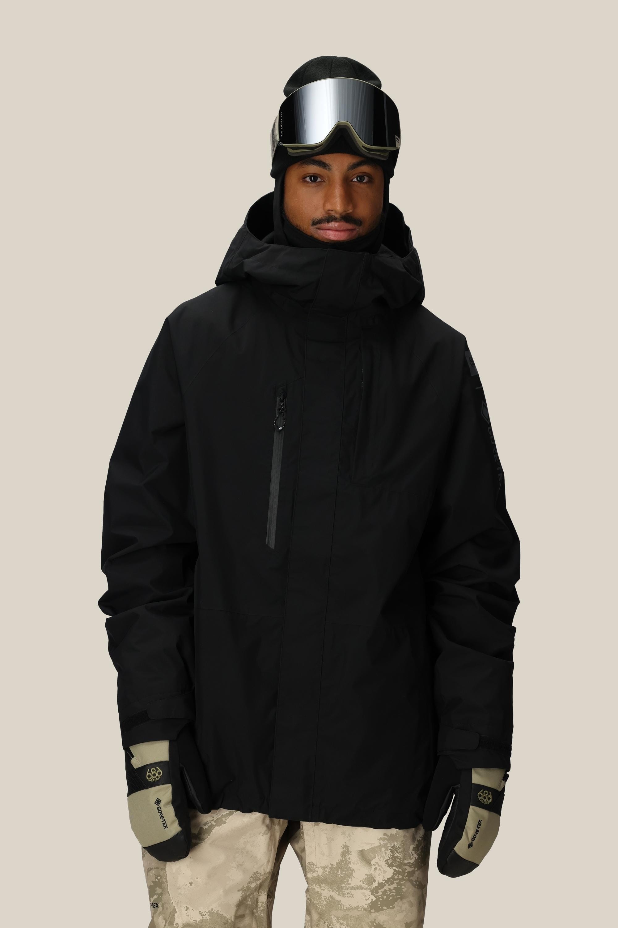 A person wearing the 686 Men’s GORE-TEX® Core Insulated Jacket in black, along with beige patterned snow pants, tan gloves, and ski goggles stands against a plain light background.