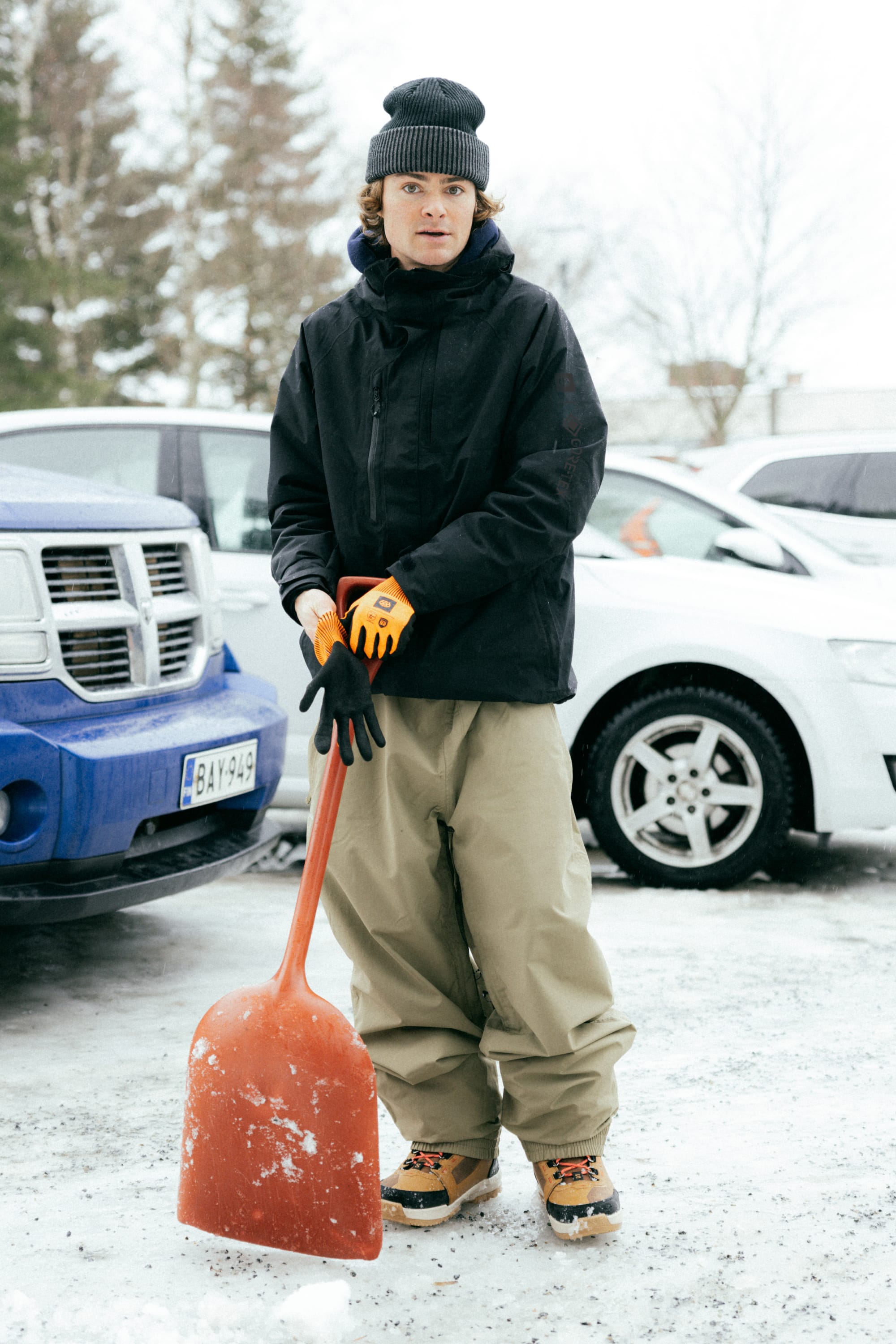 A person in winter clothing stands on snowy ground between parked cars, holding an orange snow shovel. They wear a beanie, gloves, boots, tan pants, and a black 686 Mens GORE-TEX Core Insulated Jacket. Trees are visible in the background.