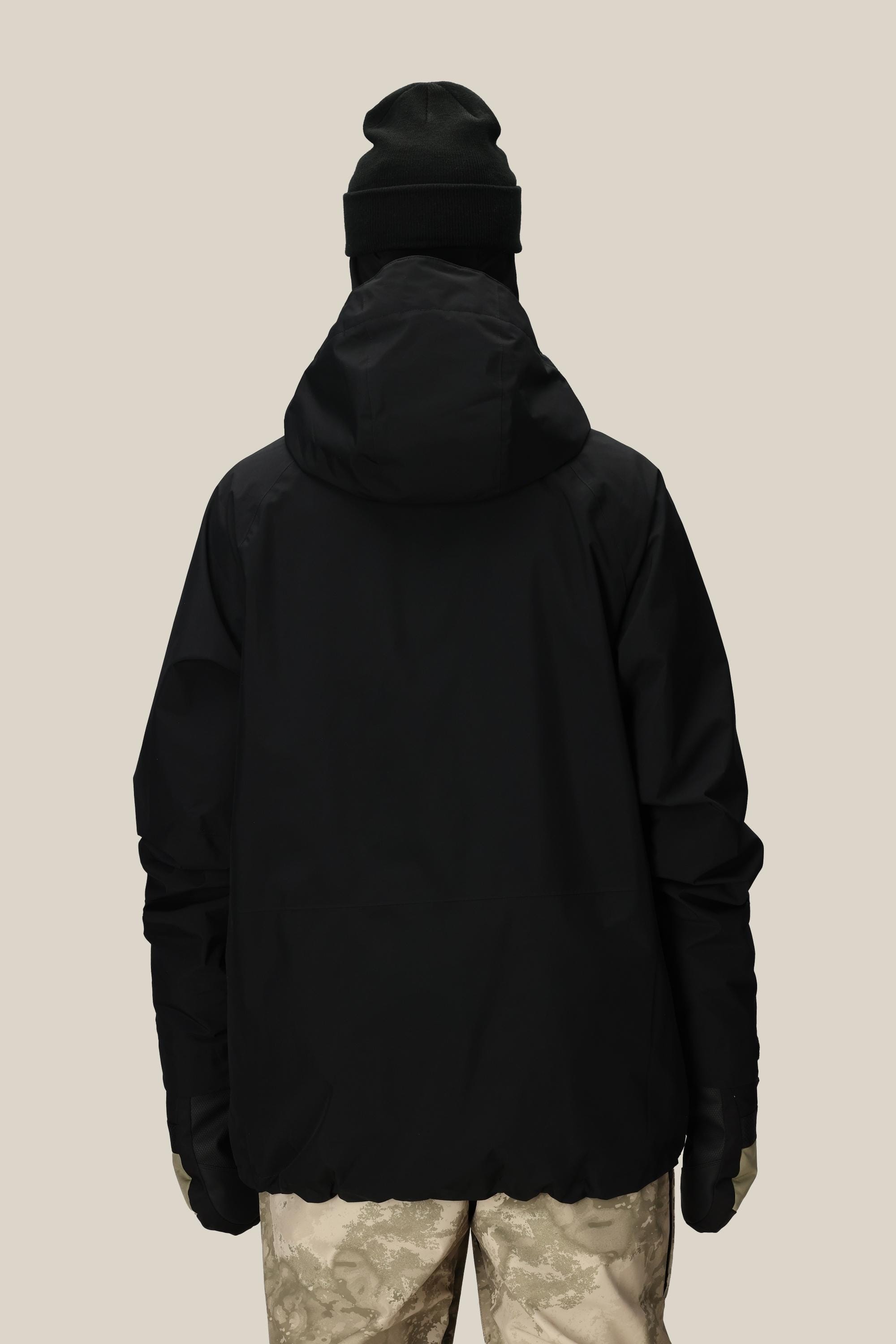 A person stands with their back to the camera in a black 686 Mens GORE-TEX Core Insulated Jacket, black beanie, black gloves, and light camo pants against a plain beige background.