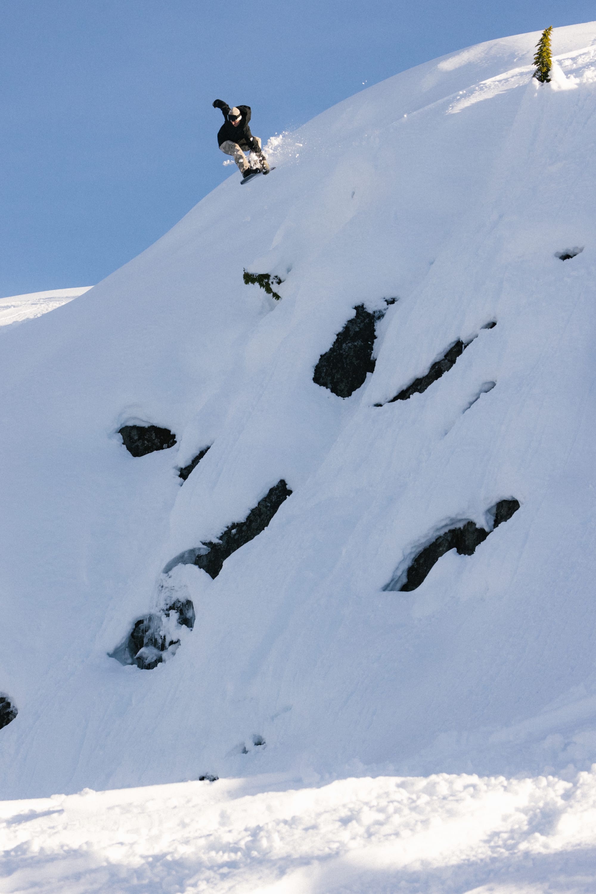 A snowboarder in a 686 Mens GORE-TEX Core Insulated Jacket rides down a steep, snowy slope dotted with rocks beneath a clear blue sky.