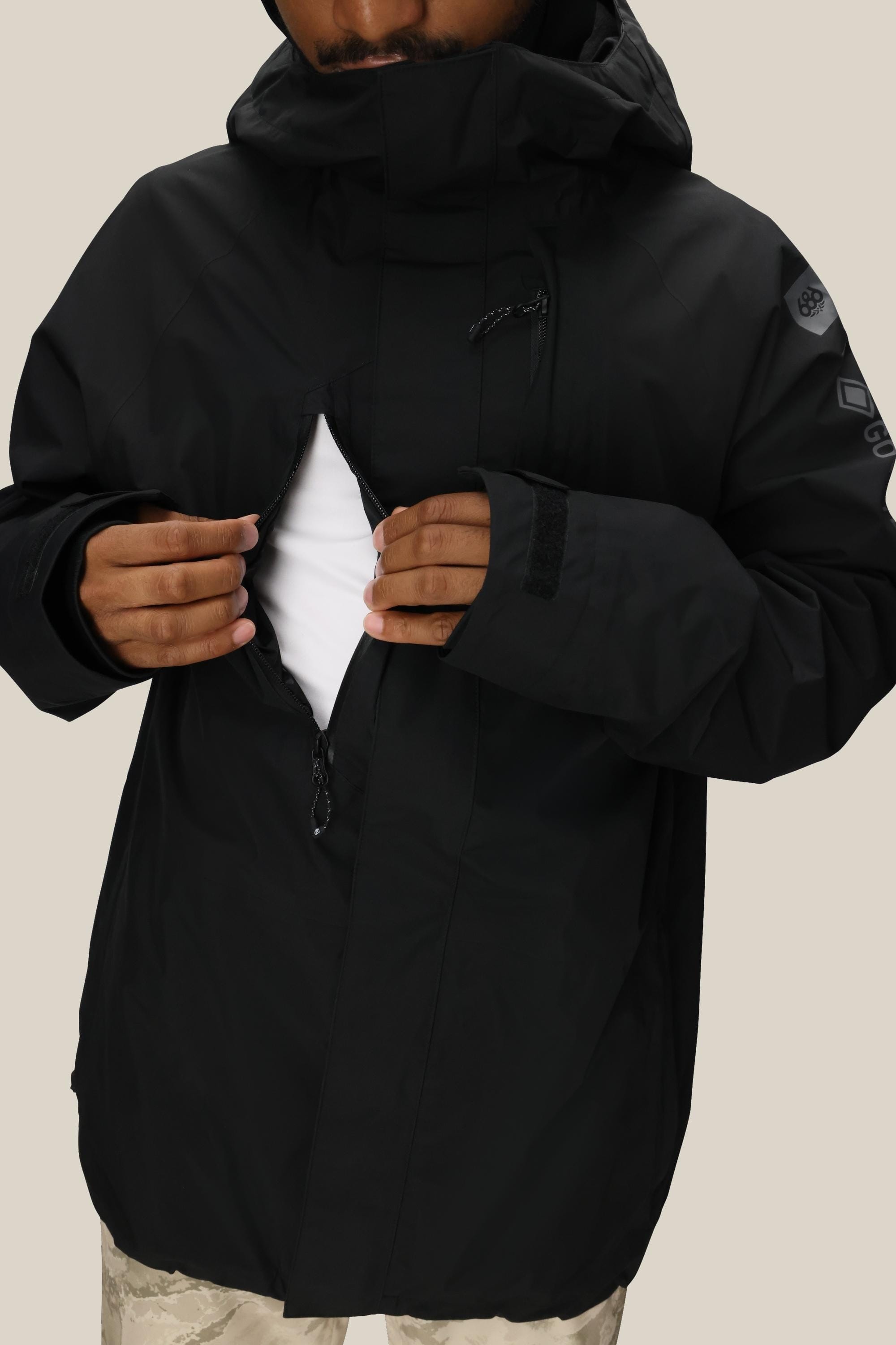 A person in the 686 Mens GORE-TEX Core Insulated Jacket unzips the chest to reveal a white shirt, showing only the lower face and torso against a plain beige background.