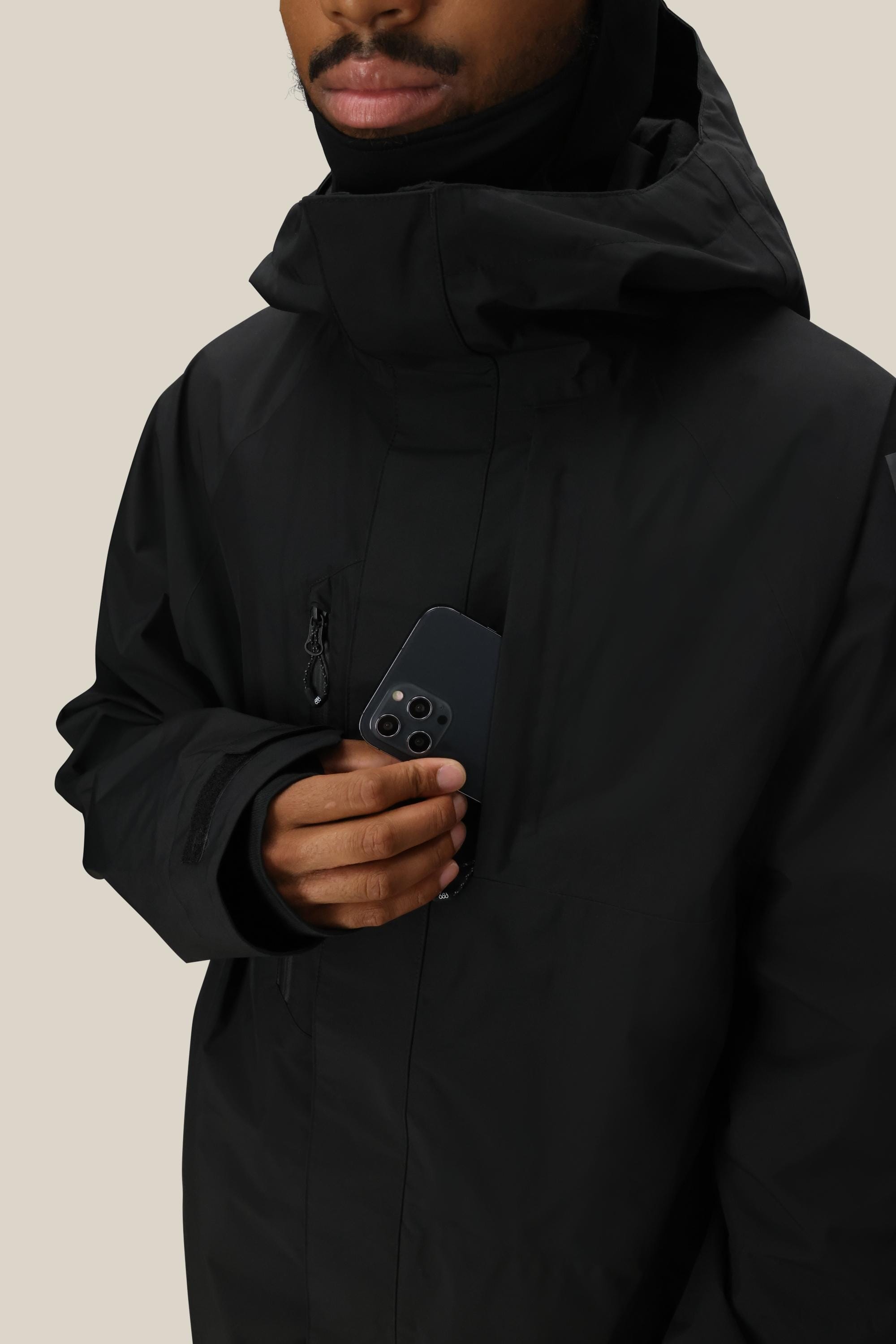 A person in a 686 Mens GORE-TEX Core Insulated Jacket holds a triple-camera smartphone, partially tucked into the chest pocket. The background is plain beige.