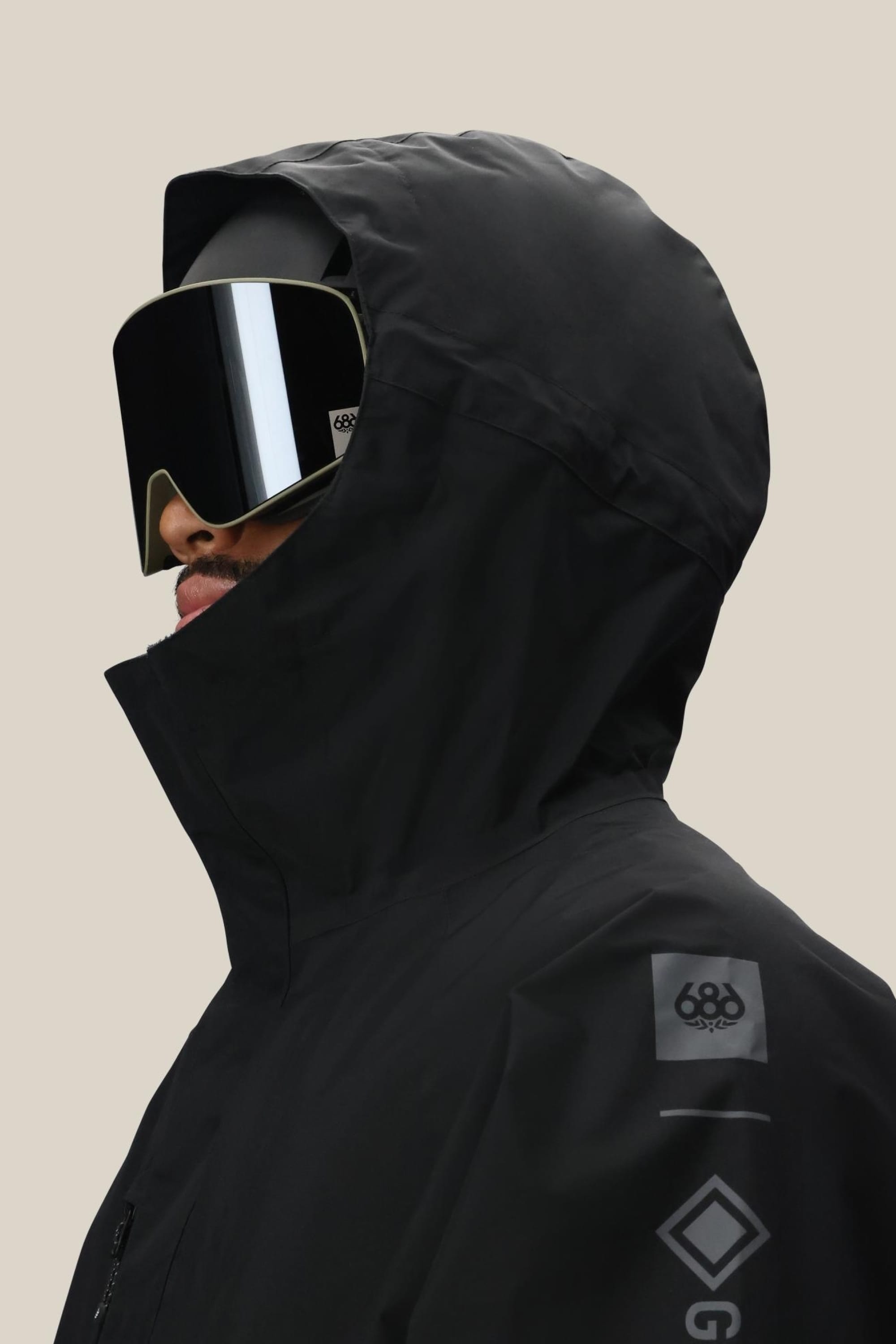 A person wears the 686 Mens GORE-TEX Core Insulated Jacket—a black waterproof polyester jacket with logo details on the shoulder—paired with a helmet and reflective ski goggles. The hood is up, standing out against a plain beige background.