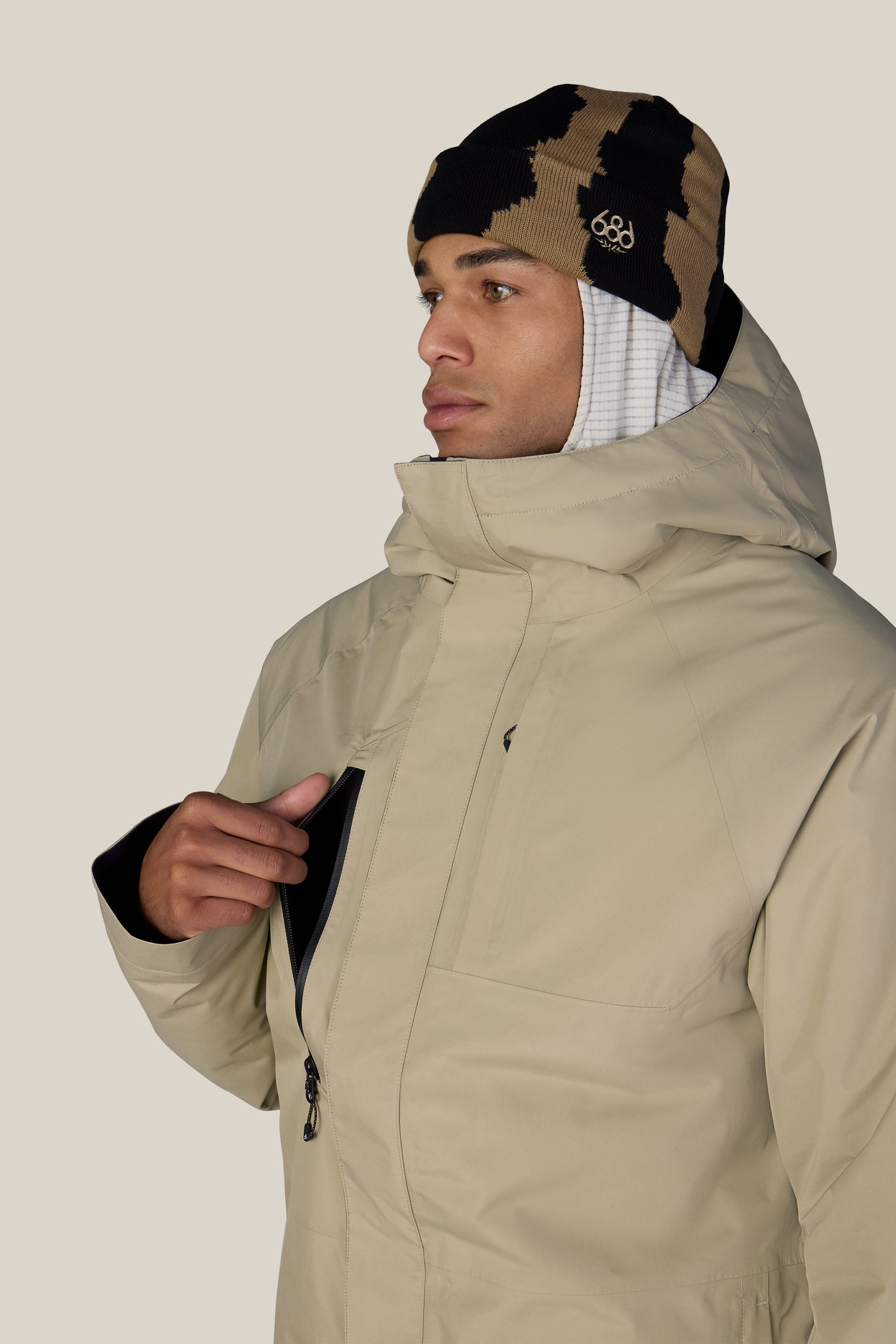A person wearing the 686 Men’s GORE-TEX® Core Insulated Jacket in beige and a patterned black and brown beanie, with a white hood underneath, opens the chest pocket of their jacket. The background is plain and light-colored.