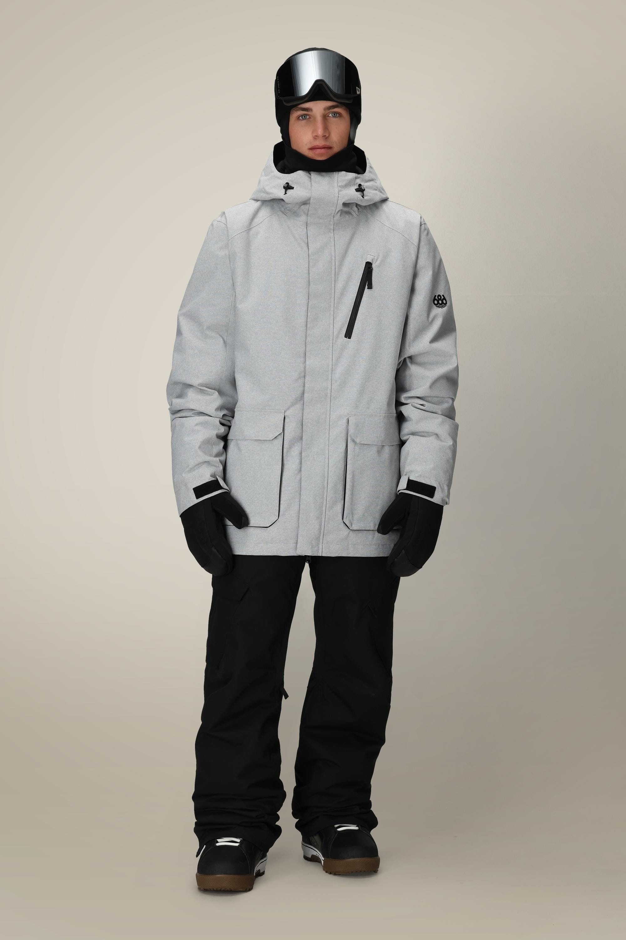 A man wearing the 686 Men's Nightline™ Insulated Jacket in light gray, black snow pants and gloves, plus a black ski helmet with goggles, stands against a plain beige background.