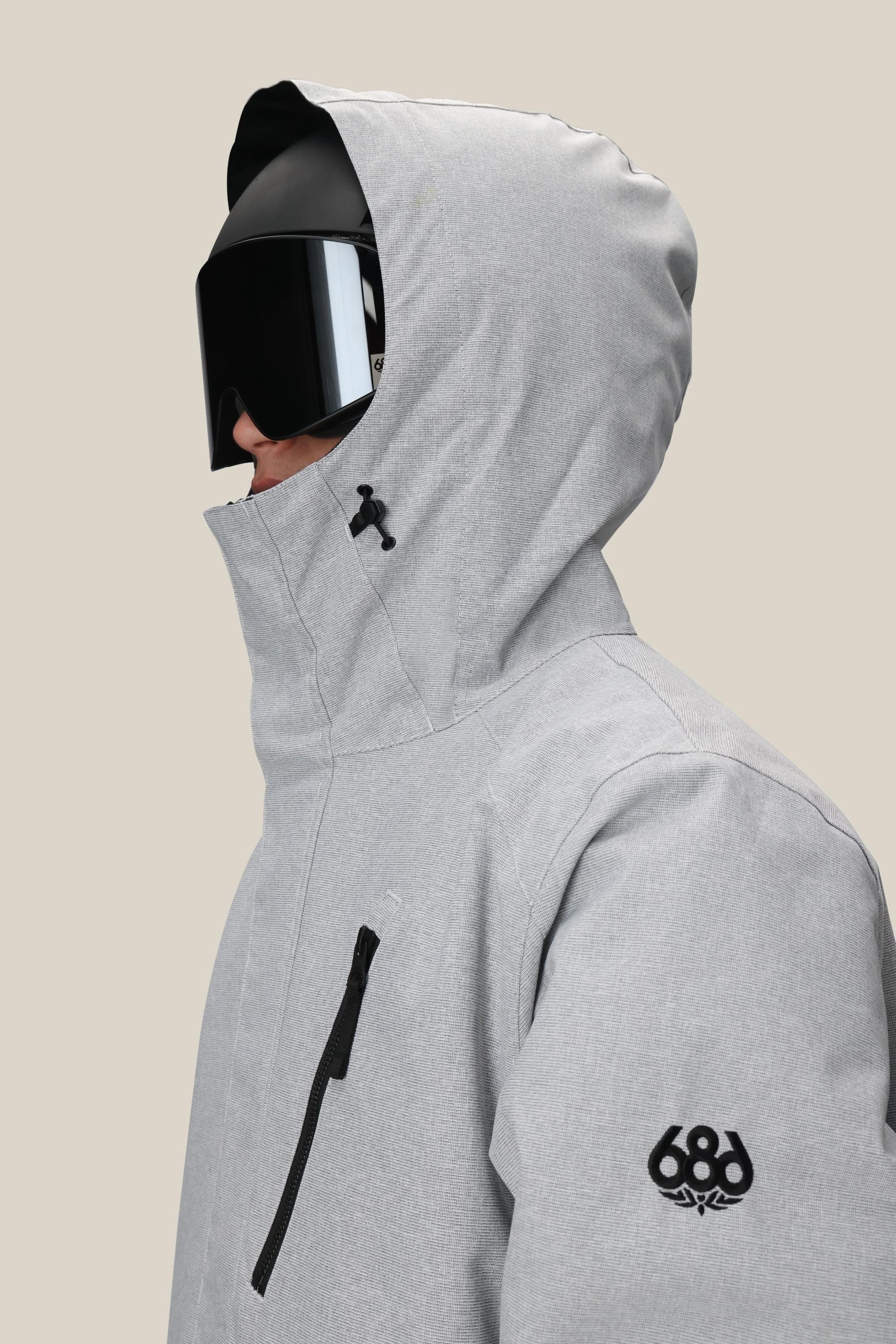 A person in profile wears the 686 Men's Nightline™ Insulated Jacket in light gray, featuring a black sleeve logo, chest zipper pocket, and large black ski goggles, set against a plain beige background.