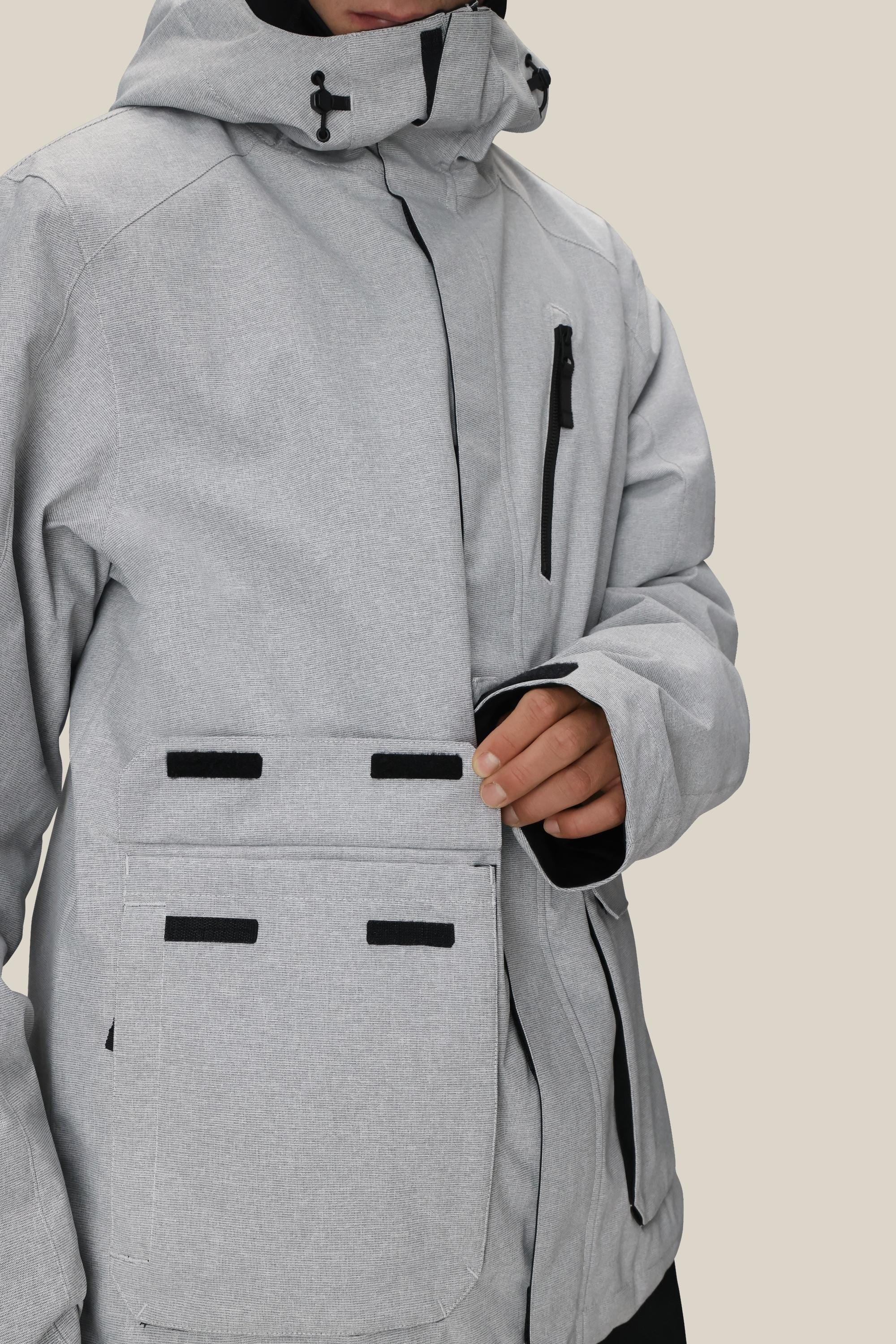 A man fastens the velcro flap on a large front pocket of the 686 Men's Nightline™ Insulated Jacket, featuring a light gray color with black zipper and velcro accents by 686.