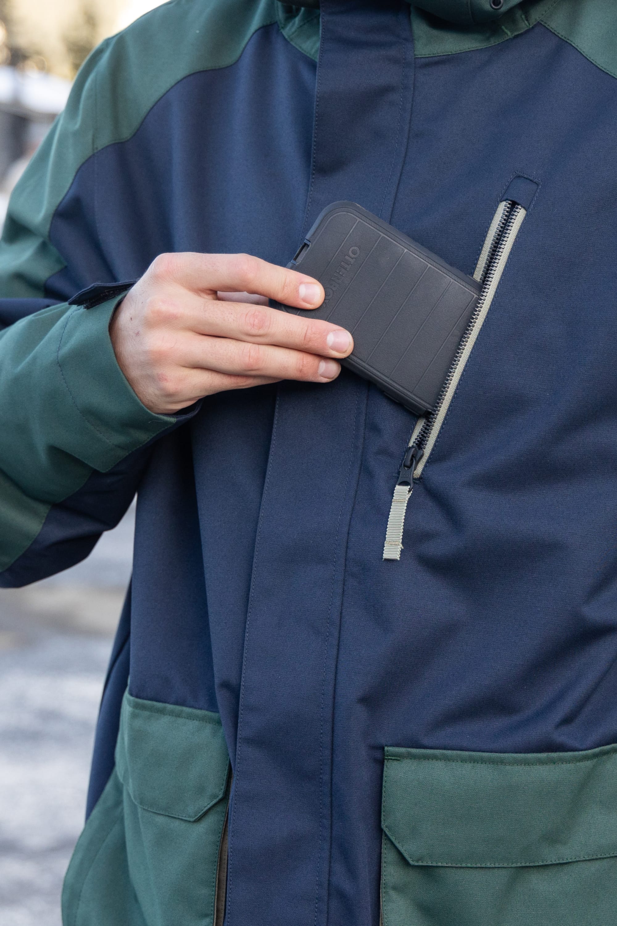 A man wearing the 686 Men's Nightline™ Insulated Jacket in green and navy places a black wallet into its zippered chest pocket.