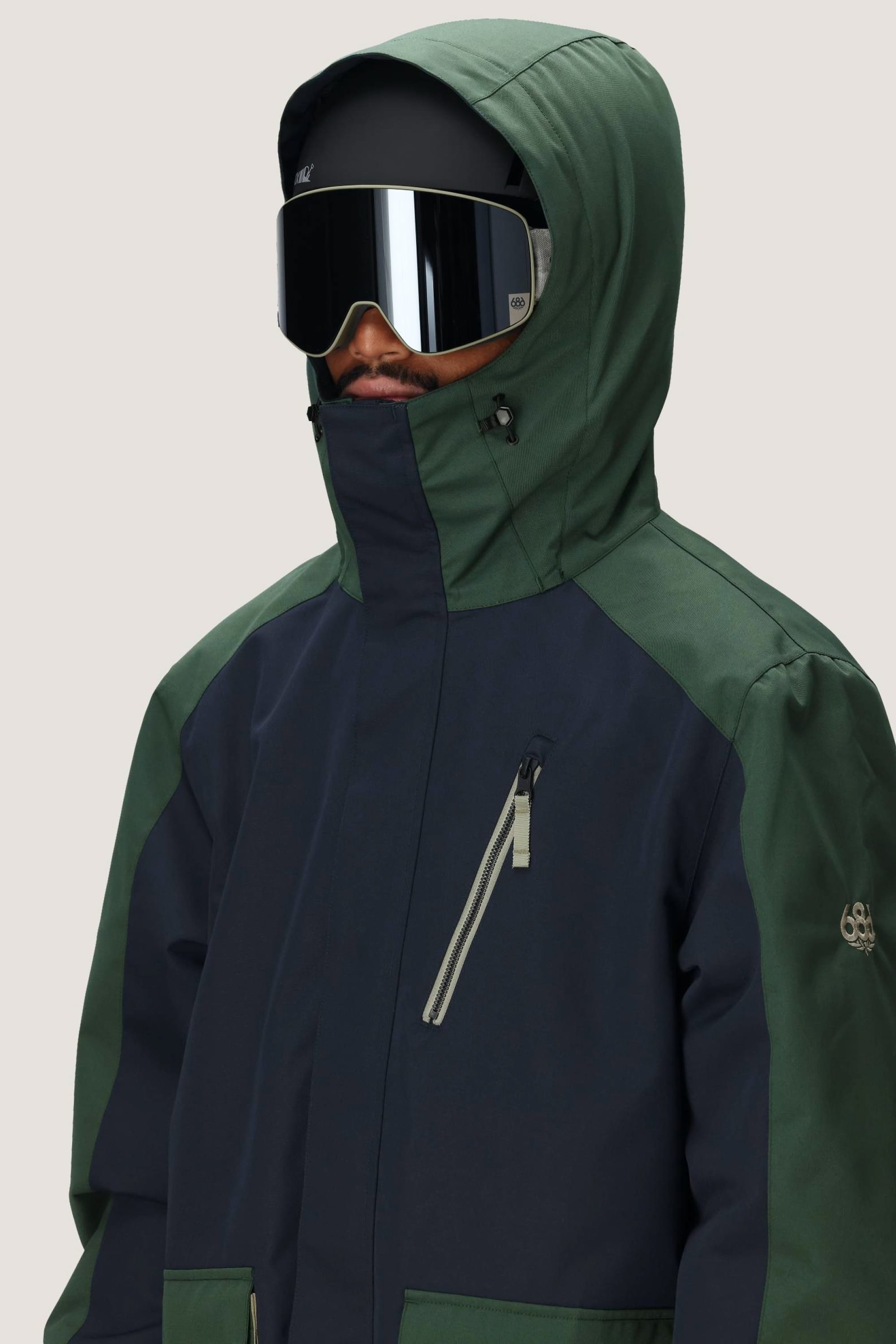 A person wears the 686 Men's Nightline™ Insulated Jacket in green and dark blue, paired with large reflective ski goggles and a black helmet. The hood is up and a plain light gray background highlights the stylish outerwear from 686.