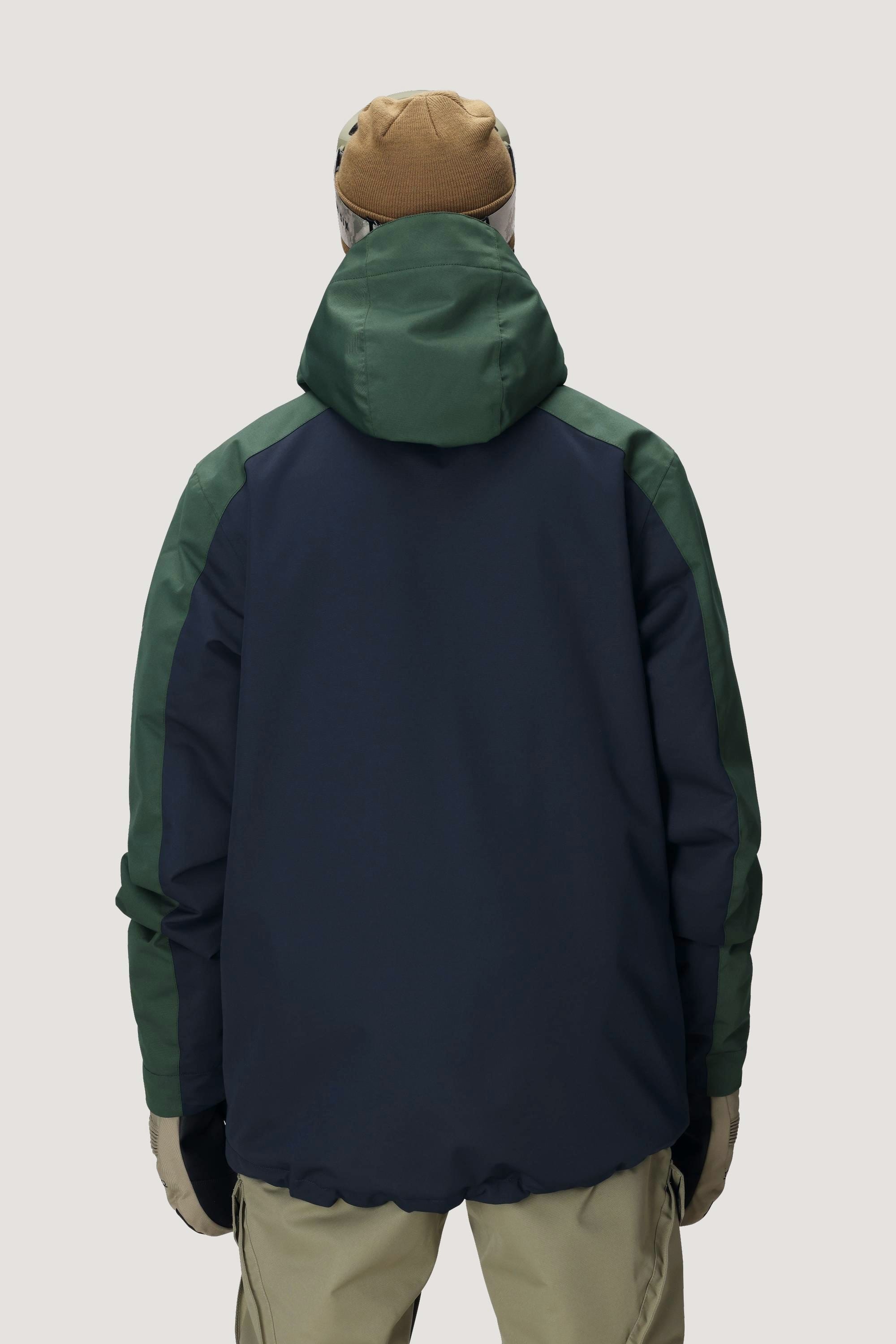 Wearing a brown beanie and the 686 Men's Nightline™ Insulated Jacket—featuring dark green sleeves, navy blue back, and hood—with beige pants, a person stands facing away from the camera against a light gray background.