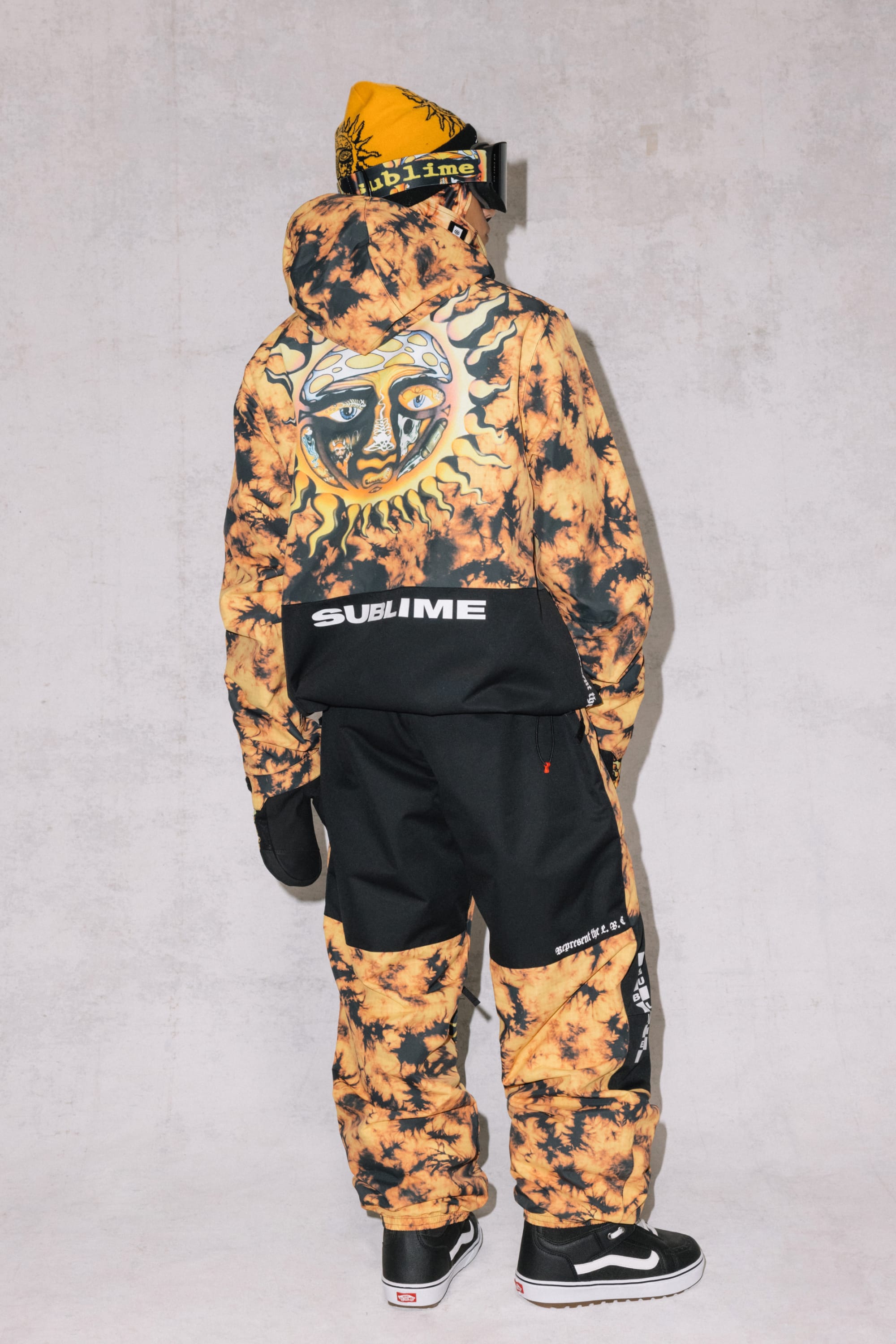 A person wears the 686 Mens Sublime Shell Anorak—yellow and black tie-dye with a sun face graphic and “Sublime” text on the back—from 686, plus a matching beanie, black gloves, and sneakers, standing against a light gray background.