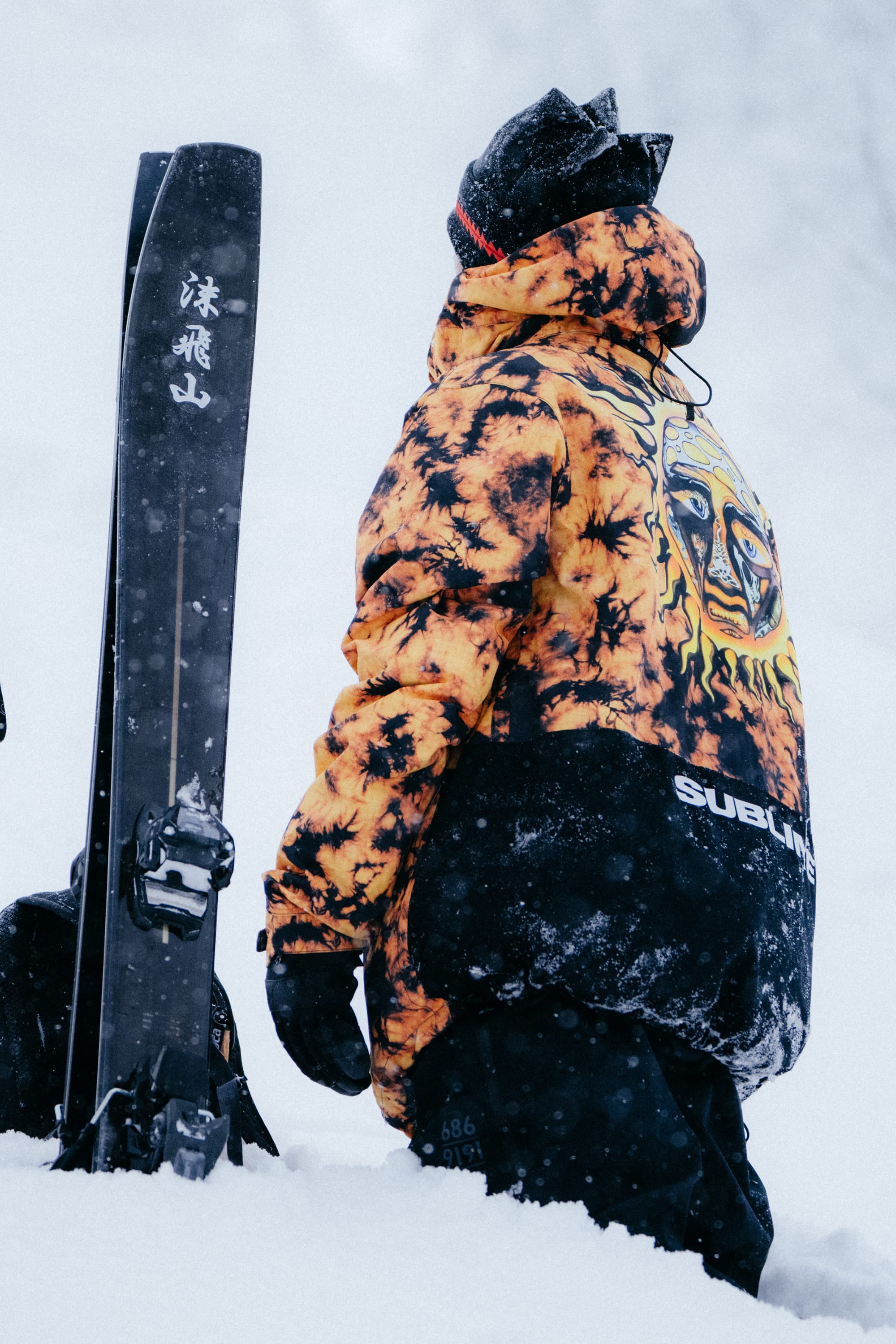 Wearing the 686 Mens Sublime Shell Anorak, a person stands in deep snow next to upright skis, looking up as snow falls, outfitted with a helmet, goggles, and gloves.