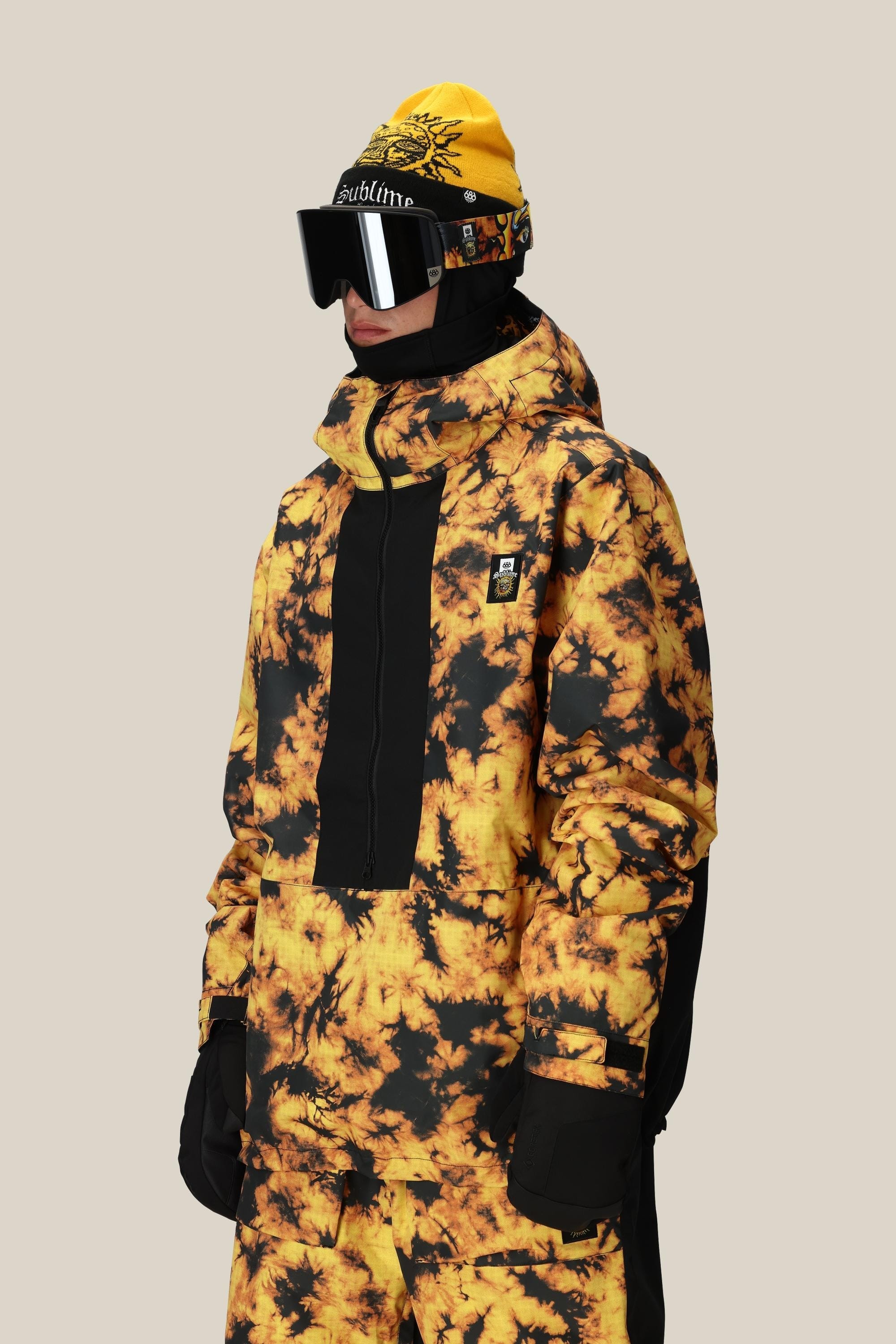 A person wears the 686 Mens Sublime Shell Anorak by 686—a yellow and black tie-dye polyester waterproof jacket—paired with black gloves, ski goggles, and a yellow beanie with black designs, set against a plain beige background.