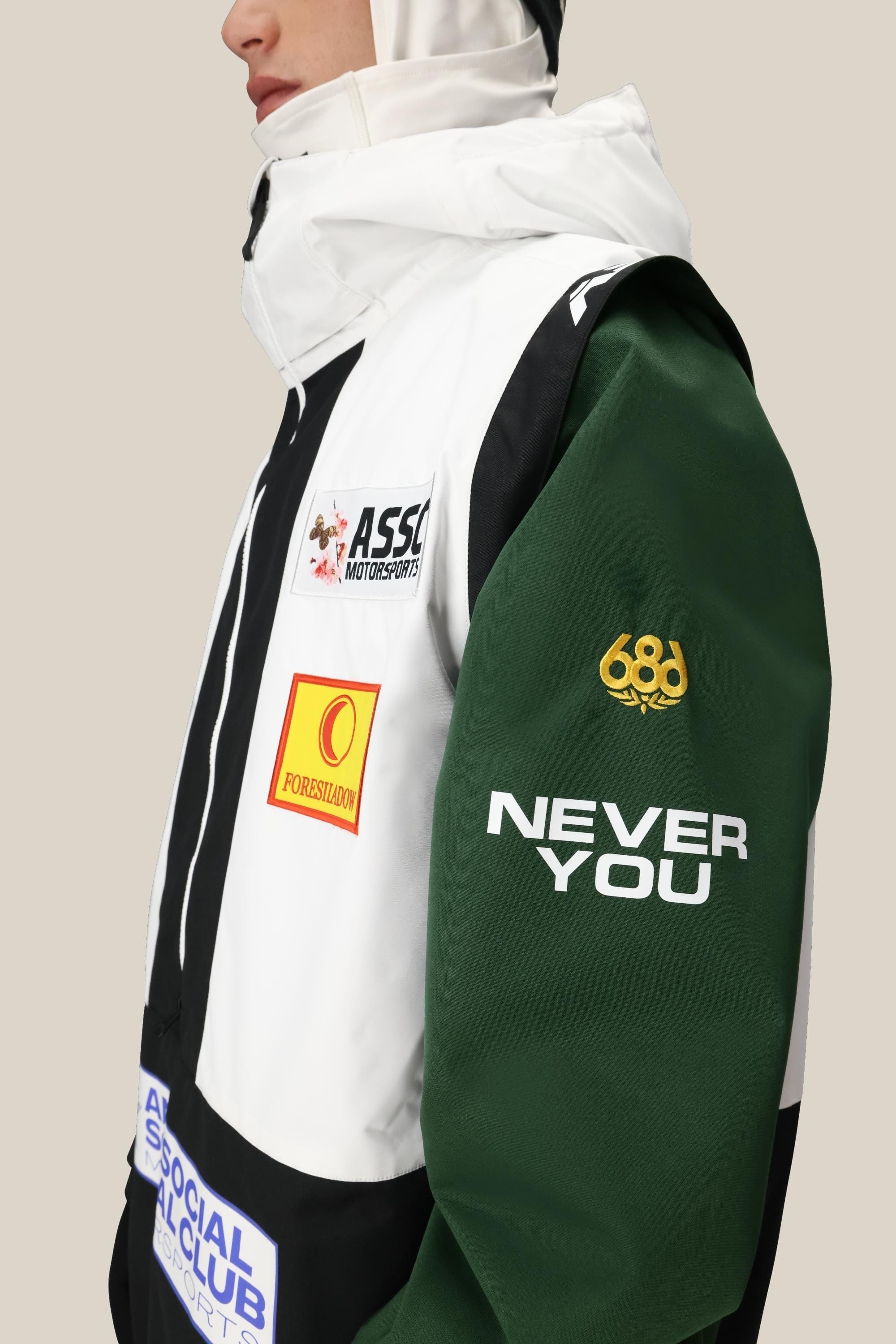 A person wears the 686 Mens ASSC F1 Shell Anorak, a color-blocked polyester waterproof jacket with green sleeves, white and black panels, racing-style logos, “NEVER YOU” on the sleeve, and assorted patches. Face partially out of frame.