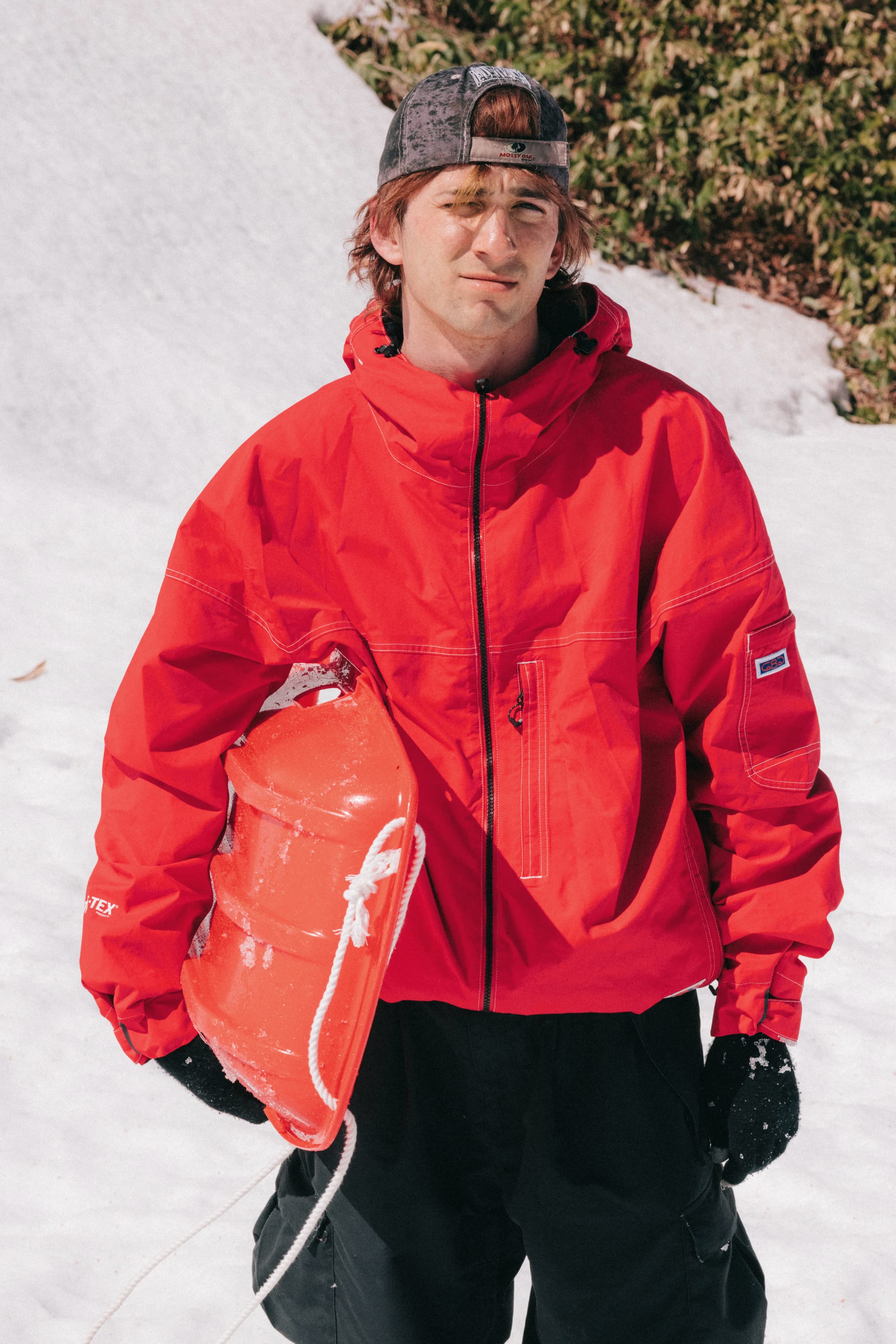 A person stands on snow wearing a red 686 Dojo® GORE-TEX Jacket, black gloves, and a backward cap, holding a red sled under one arm. Green bushes are visible in the background.